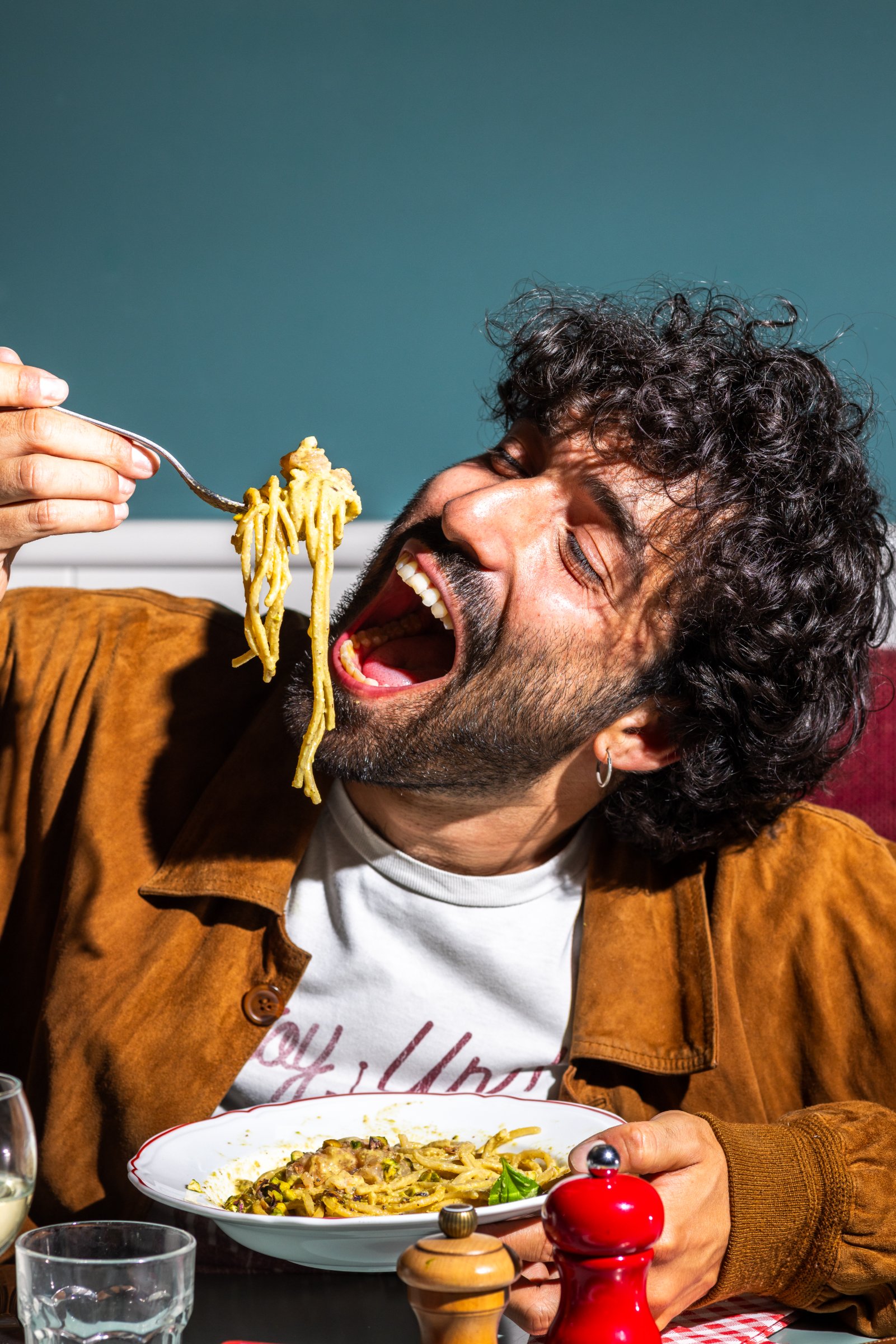 A man with curly hair enthusiastically eats a plate of pasta in a restaurant with dramatic lighting.