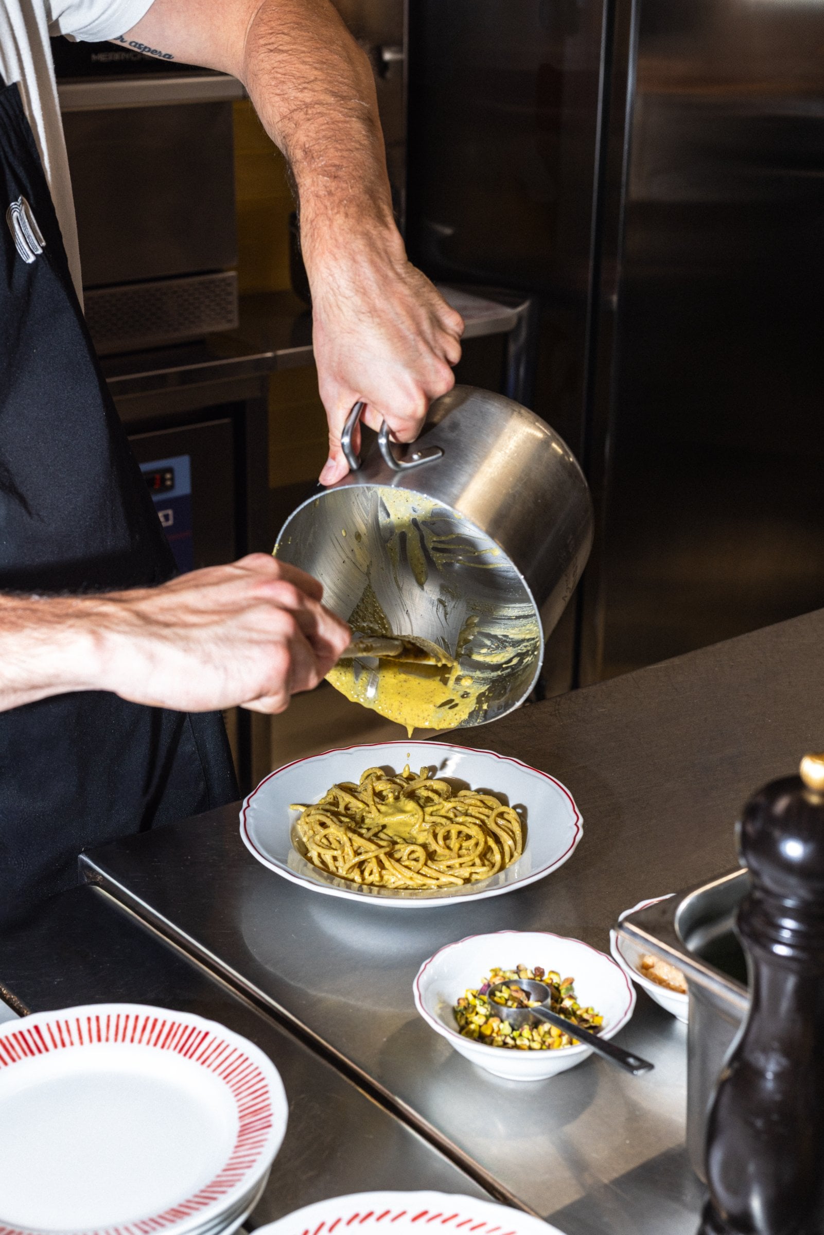 A chef pours a creamy pistachio sauce over a plate of freshly cooked pasta in a professional kitchen.