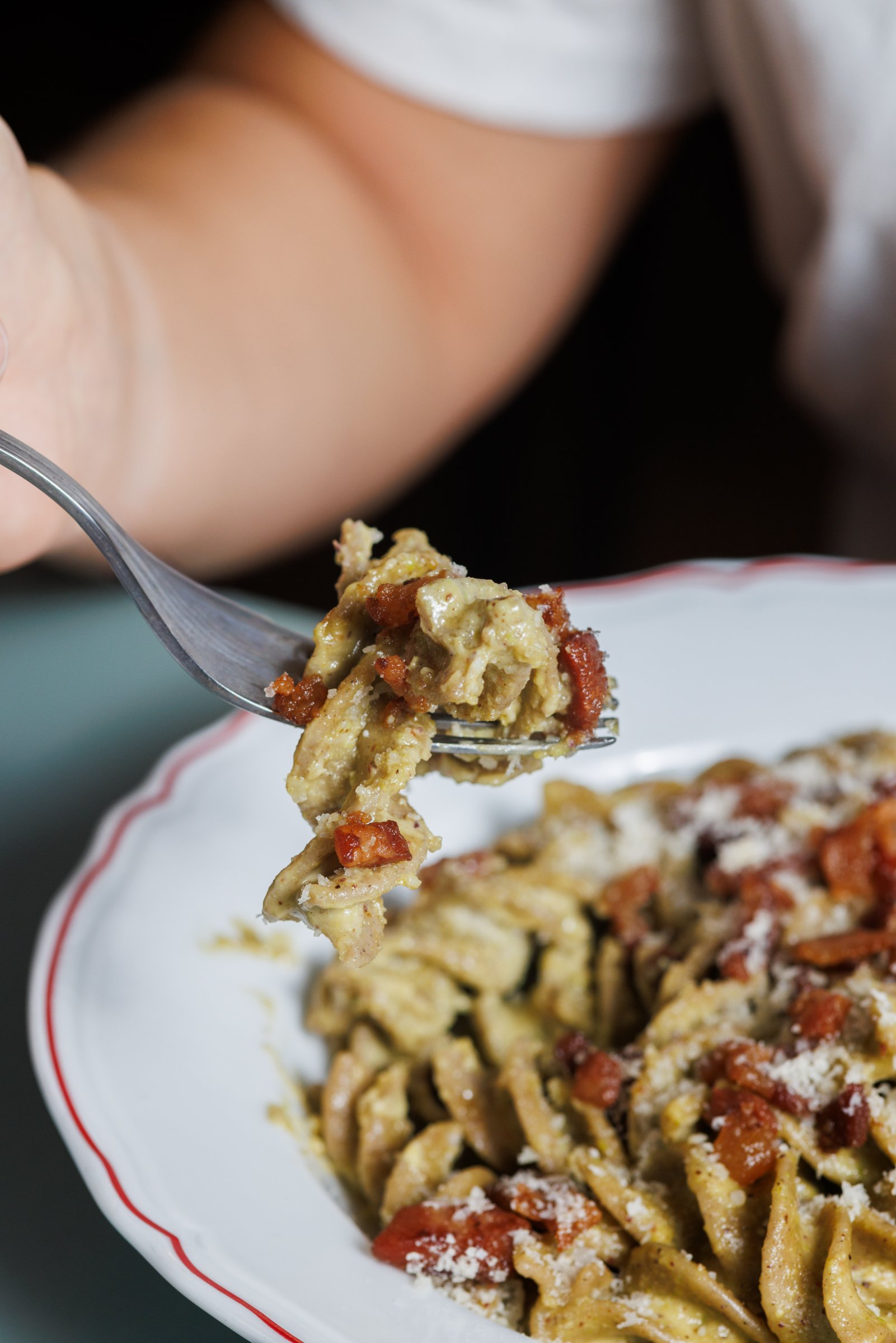 A close-up of a fork lifting a portion of creamy pasta topped with crispy bacon bits and grated cheese.