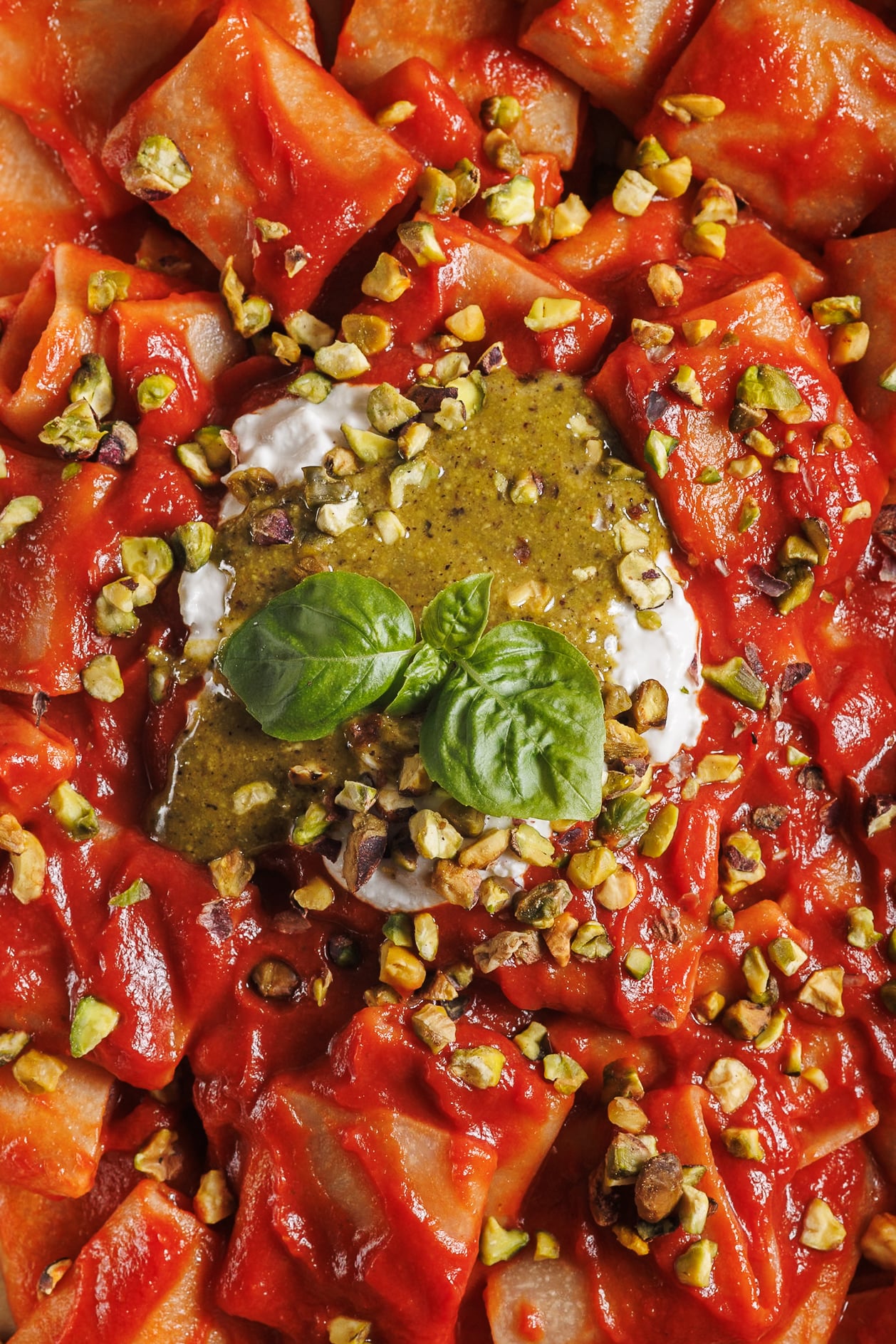 A close-up overhead shot of pasta tossed in a vibrant tomato sauce, topped with creamy burrata, pesto, and crushed pistachios.