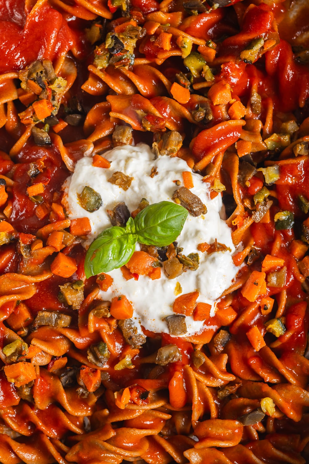 A close-up of rotini pasta tossed in a rich tomato sauce with diced vegetables and topped with a dollop of creamy ricotta cheese and fresh basil.