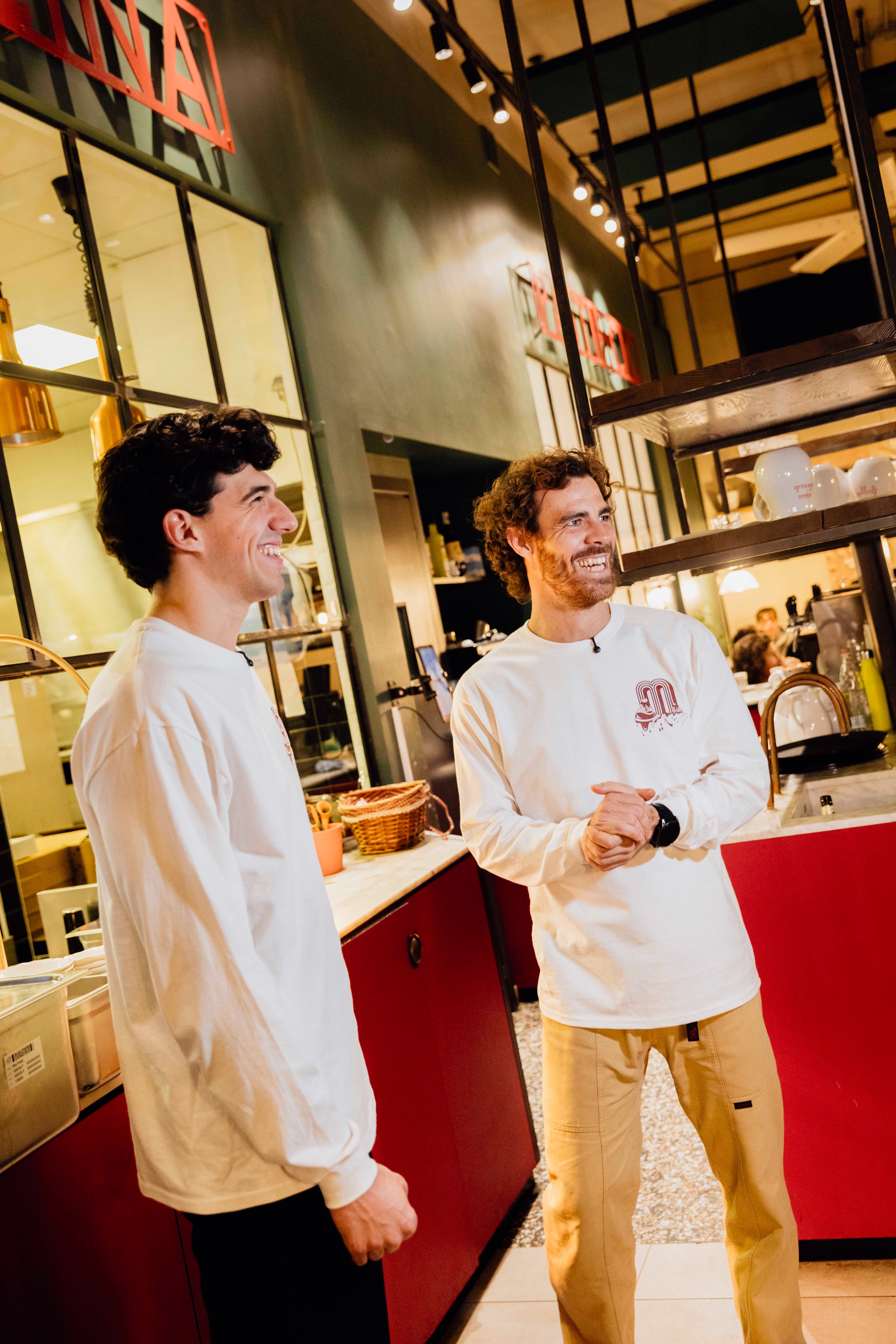 Two men in matching long-sleeved shirts stand in a modern, well-lit restaurant kitchen.