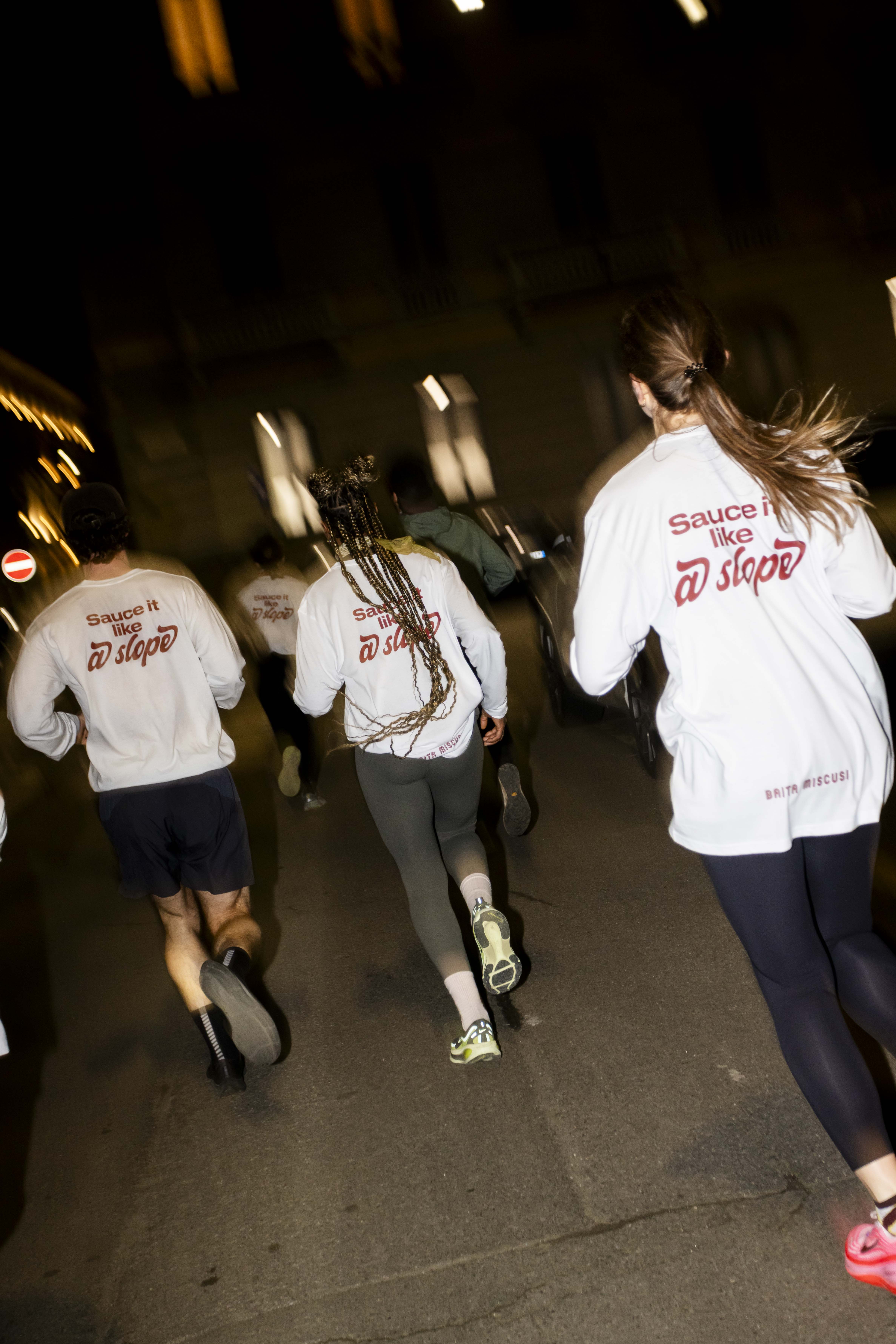 A group of people wearing branded long-sleeve shirts run together on a city street at night.