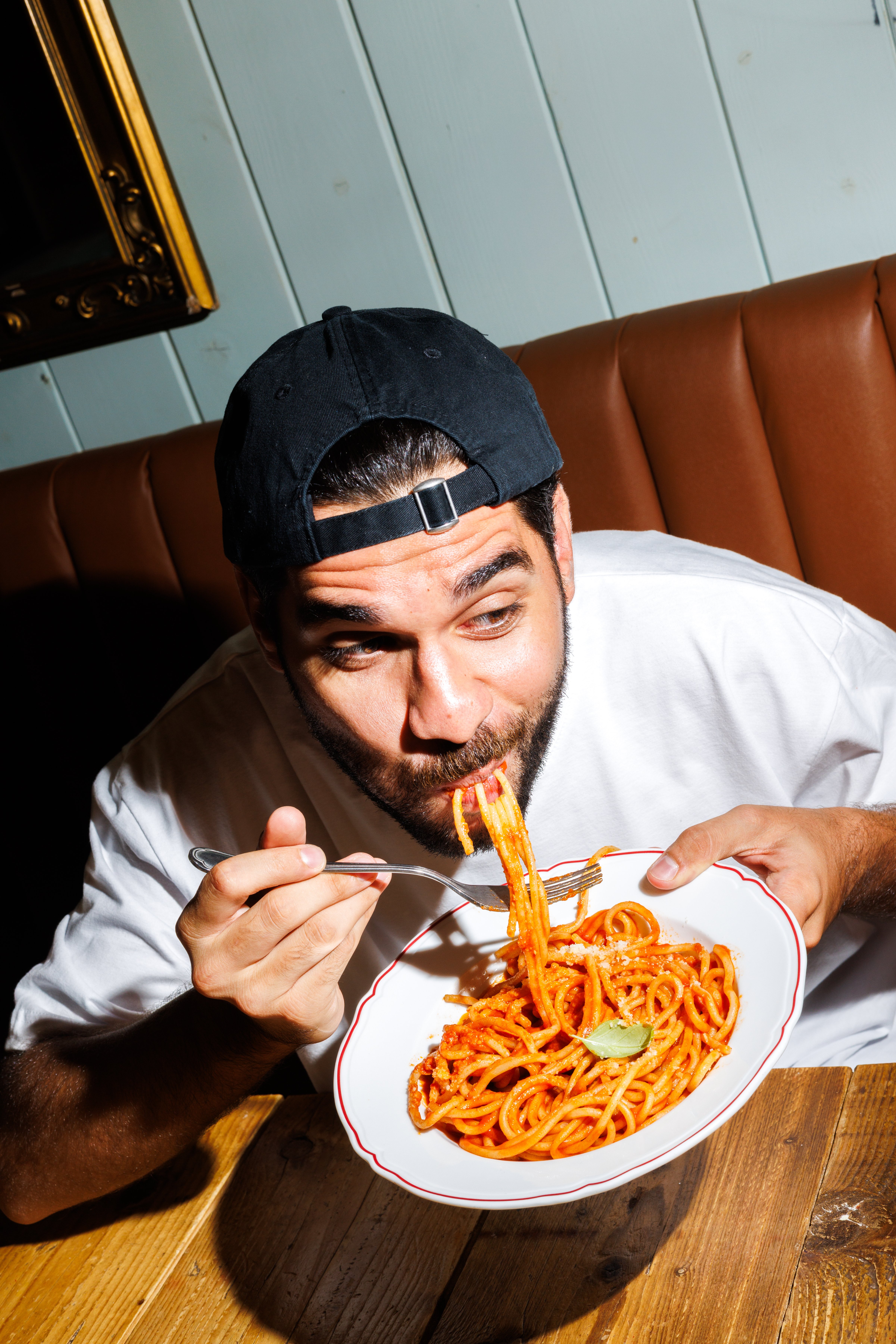 A man in a baseball cap enjoys a plate of tomato-based pasta in a dimly lit restaurant setting.