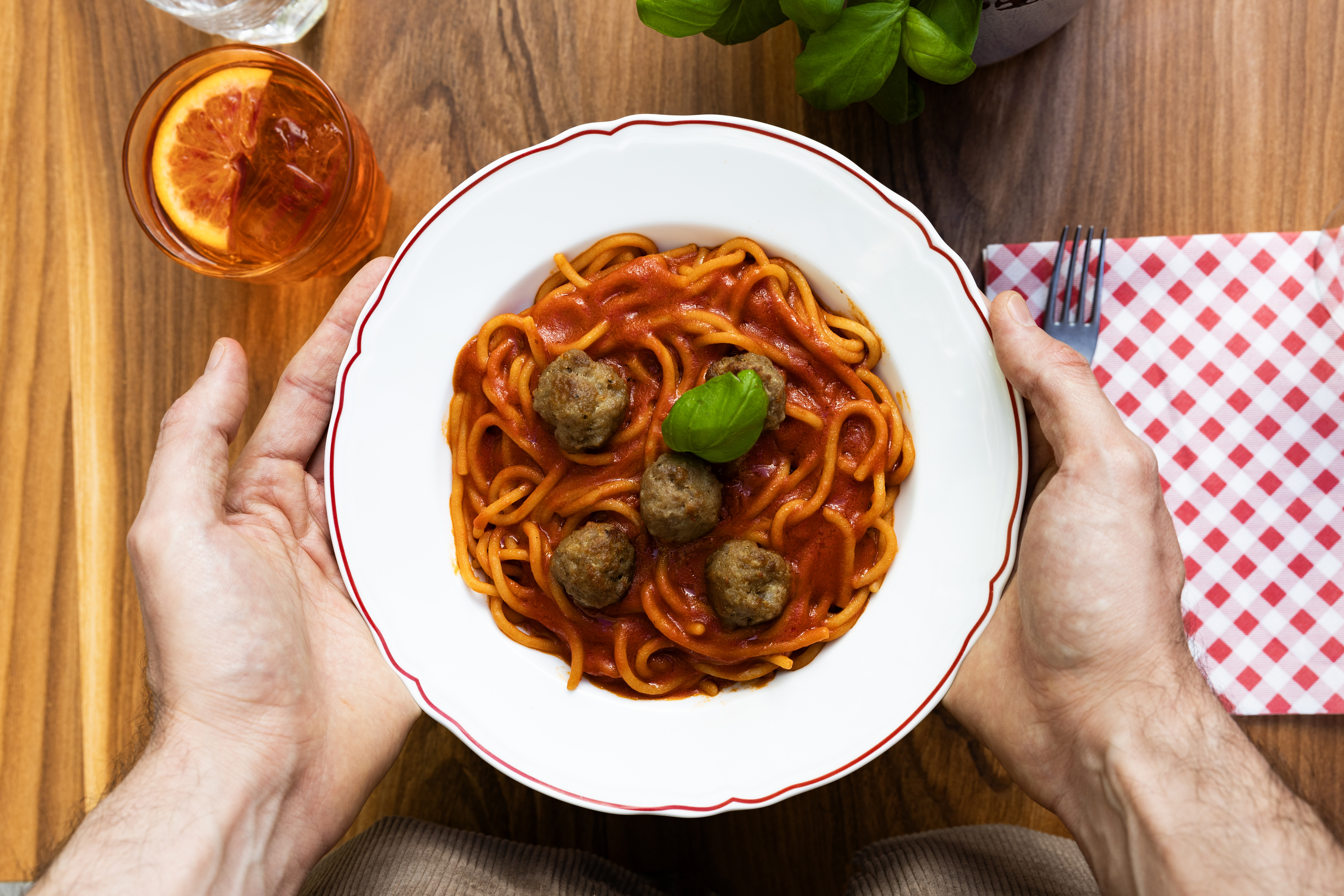 A person holds a plate of spaghetti with meatballs and a fresh basil garnish above a wooden table.
