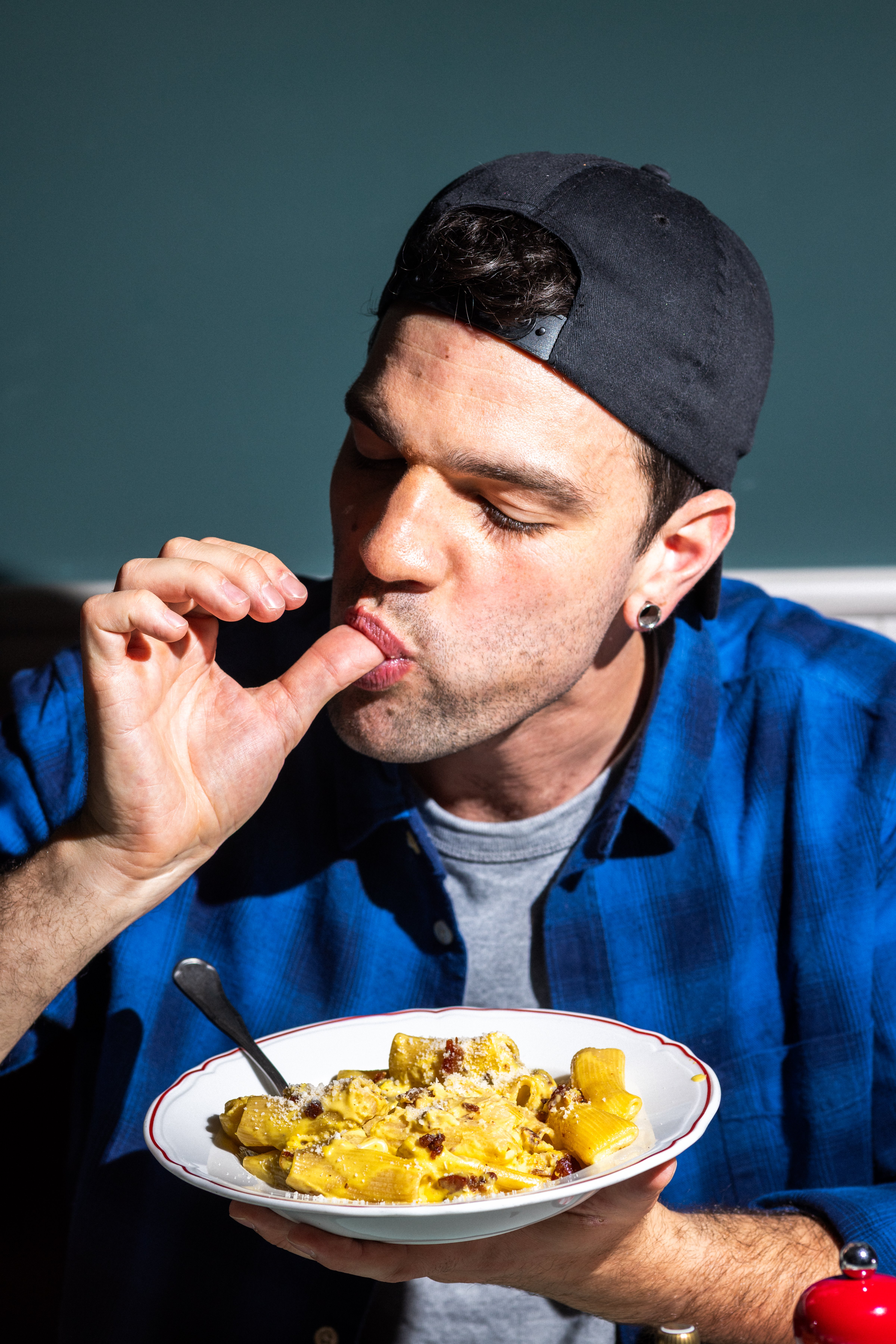 A man in a blue flannel shirt enjoys a plate of creamy pasta while dining in a dimly lit setting.