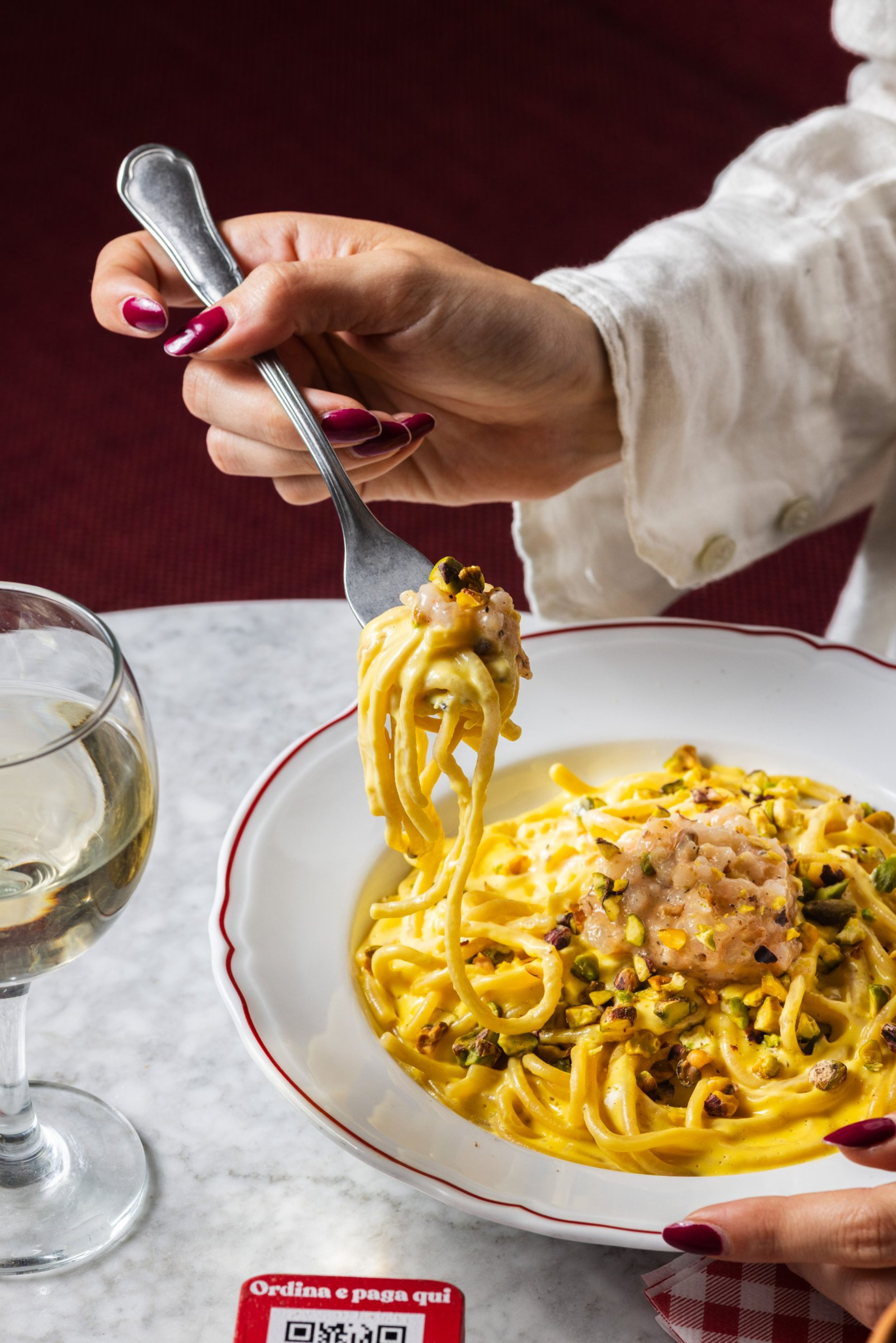 A person uses a fork to lift creamy pasta topped with crushed pistachios and raw seafood from a white plate.