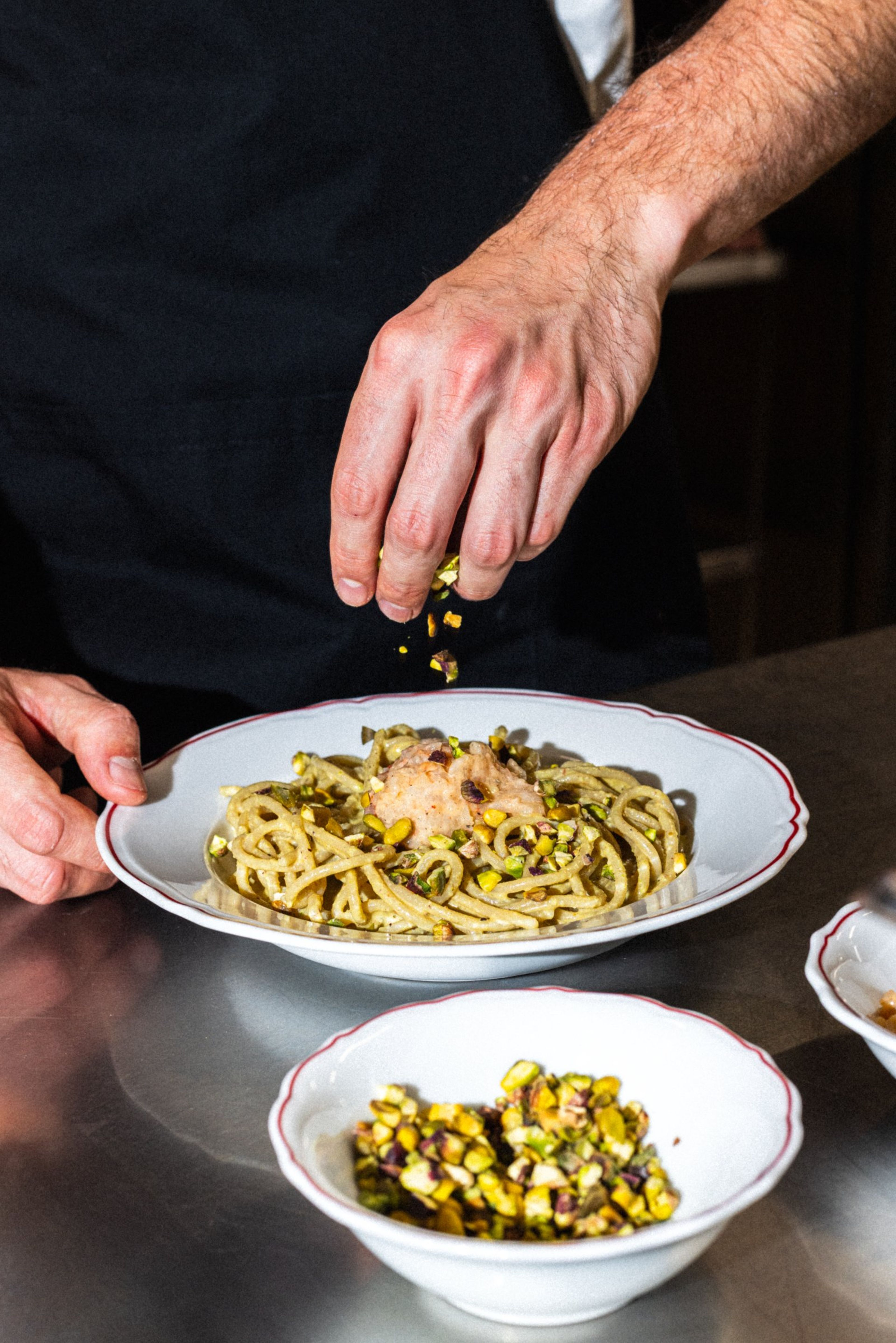 A chef sprinkles crushed pistachios over a plate of pasta in a professional kitchen setting.