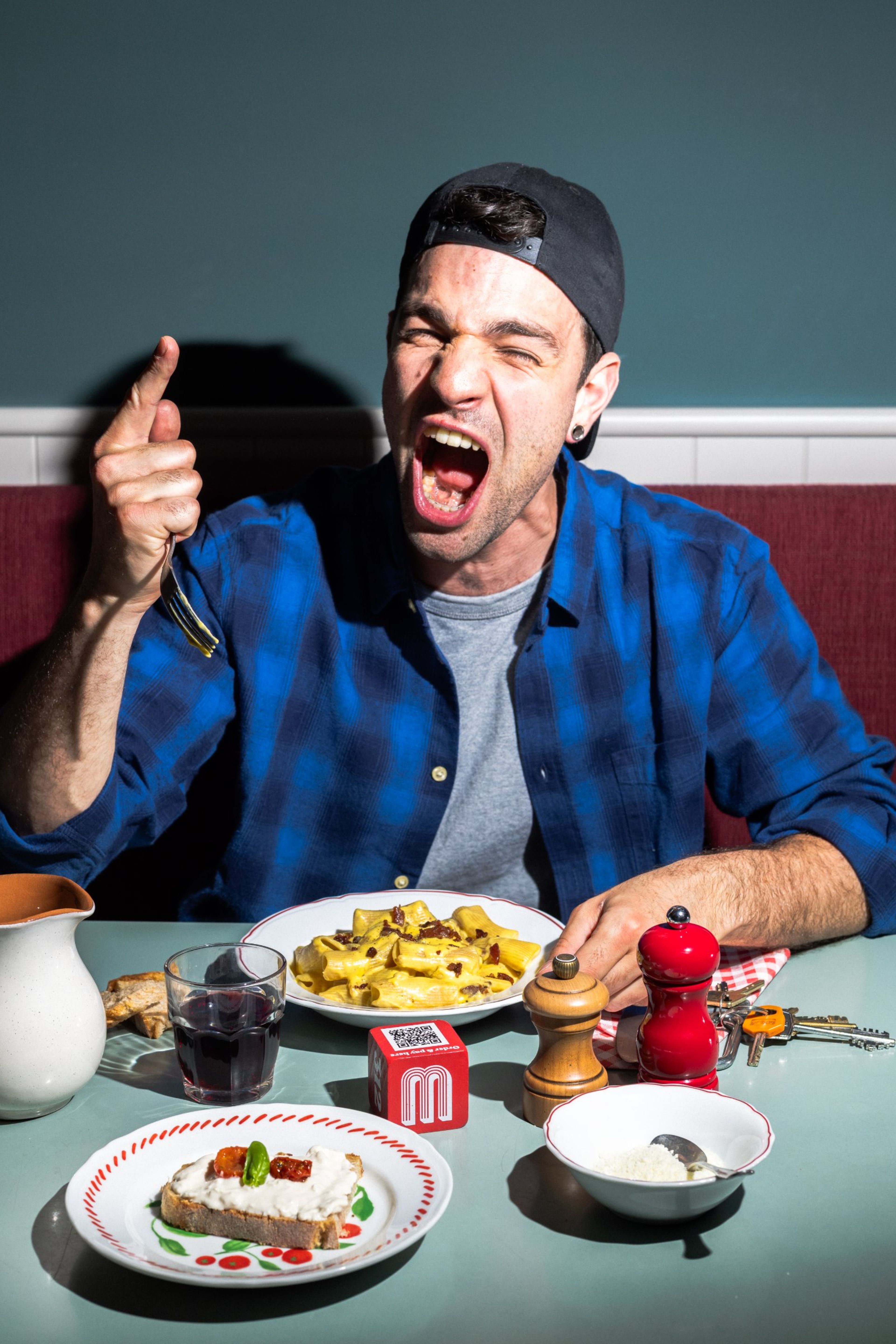 A man in a blue plaid shirt shouts excitedly while sitting at a restaurant table with pasta, bread, and wine.