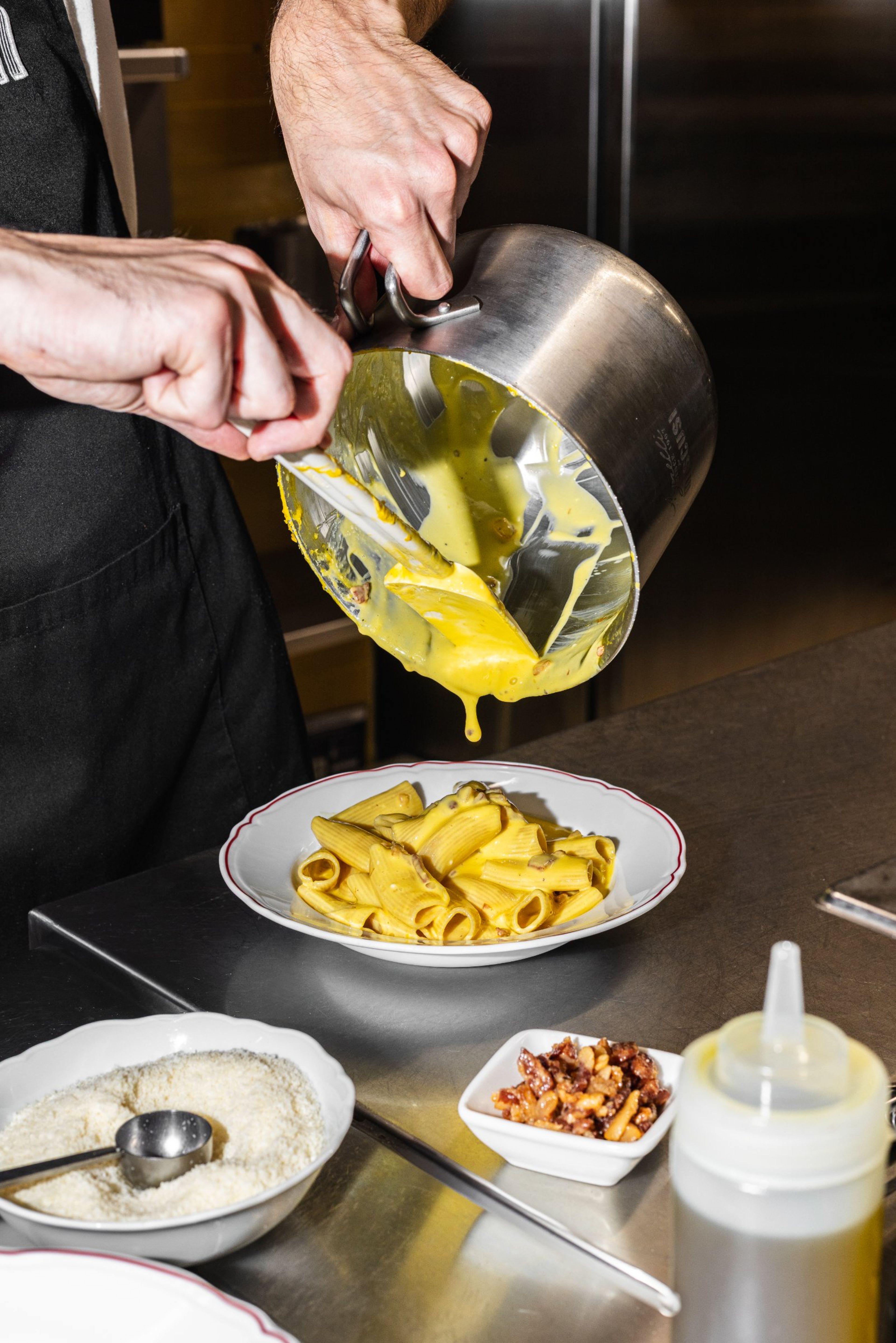 A chef pours a creamy yellow sauce over a plate of rigatoni pasta in a professional kitchen.