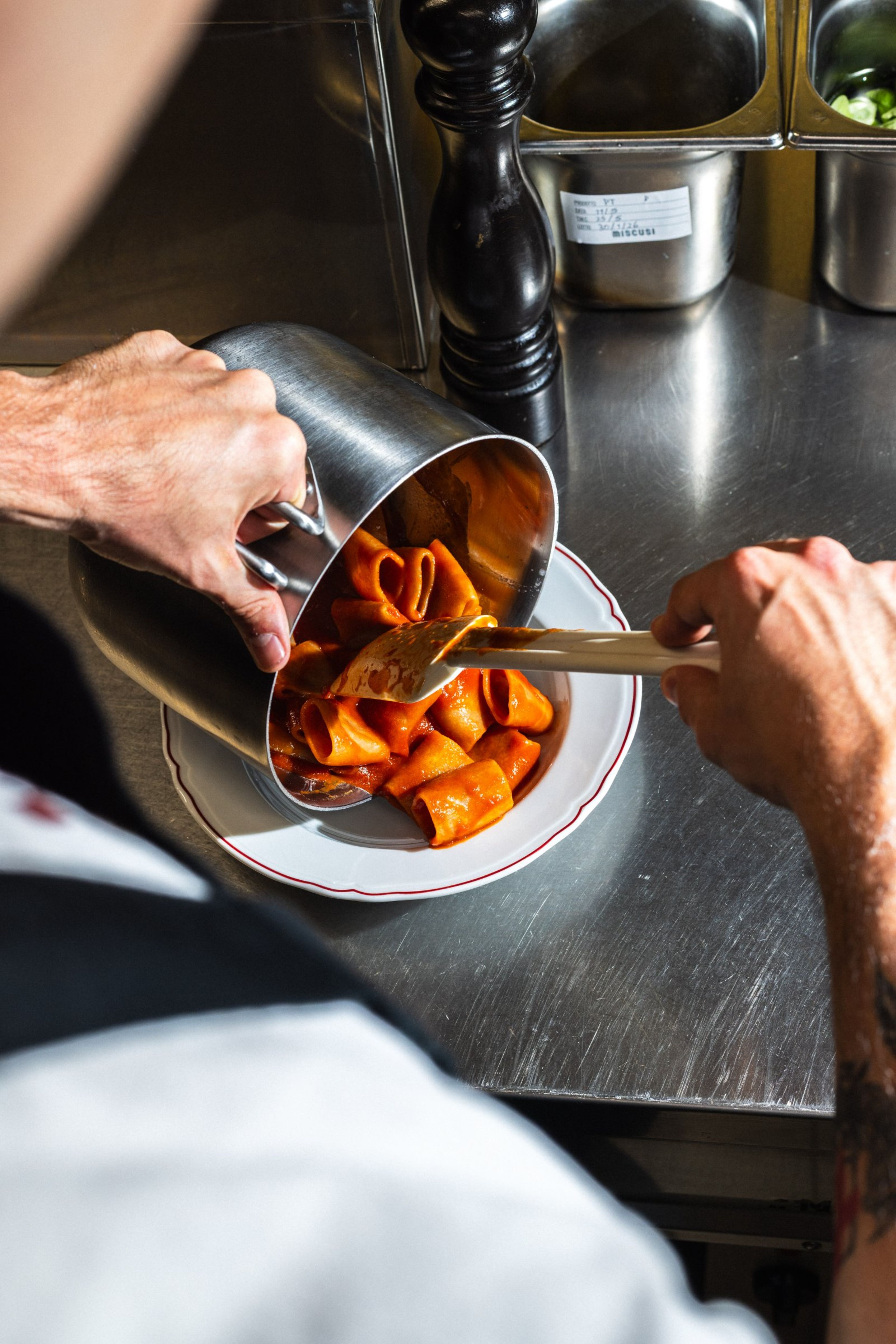 A chef uses a spatula to plate freshly cooked rigatoni pasta with tomato sauce onto a white dish in a professional kitchen.