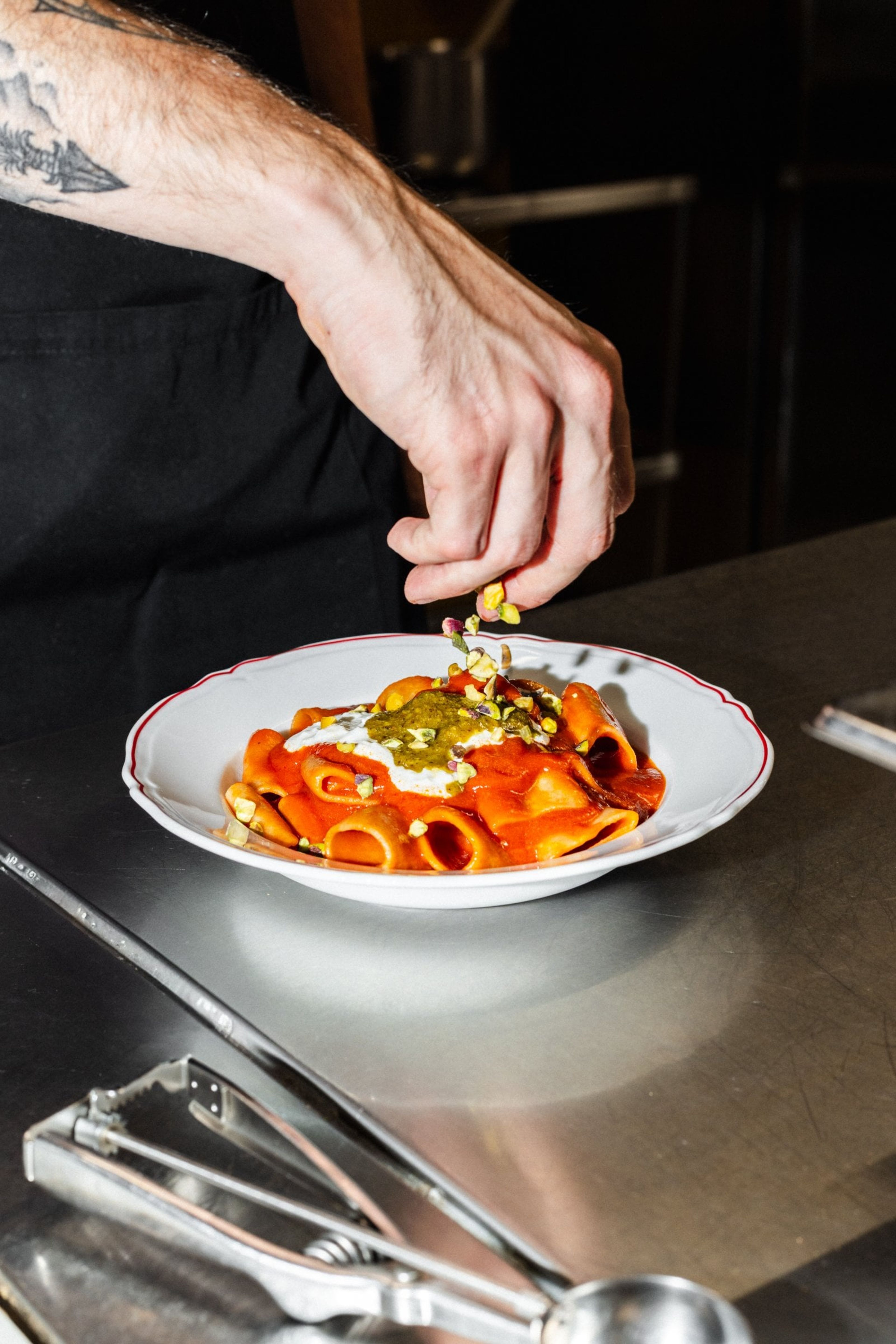 A chef sprinkles crushed pistachios over a plate of pasta topped with tomato sauce and creamy cheese.