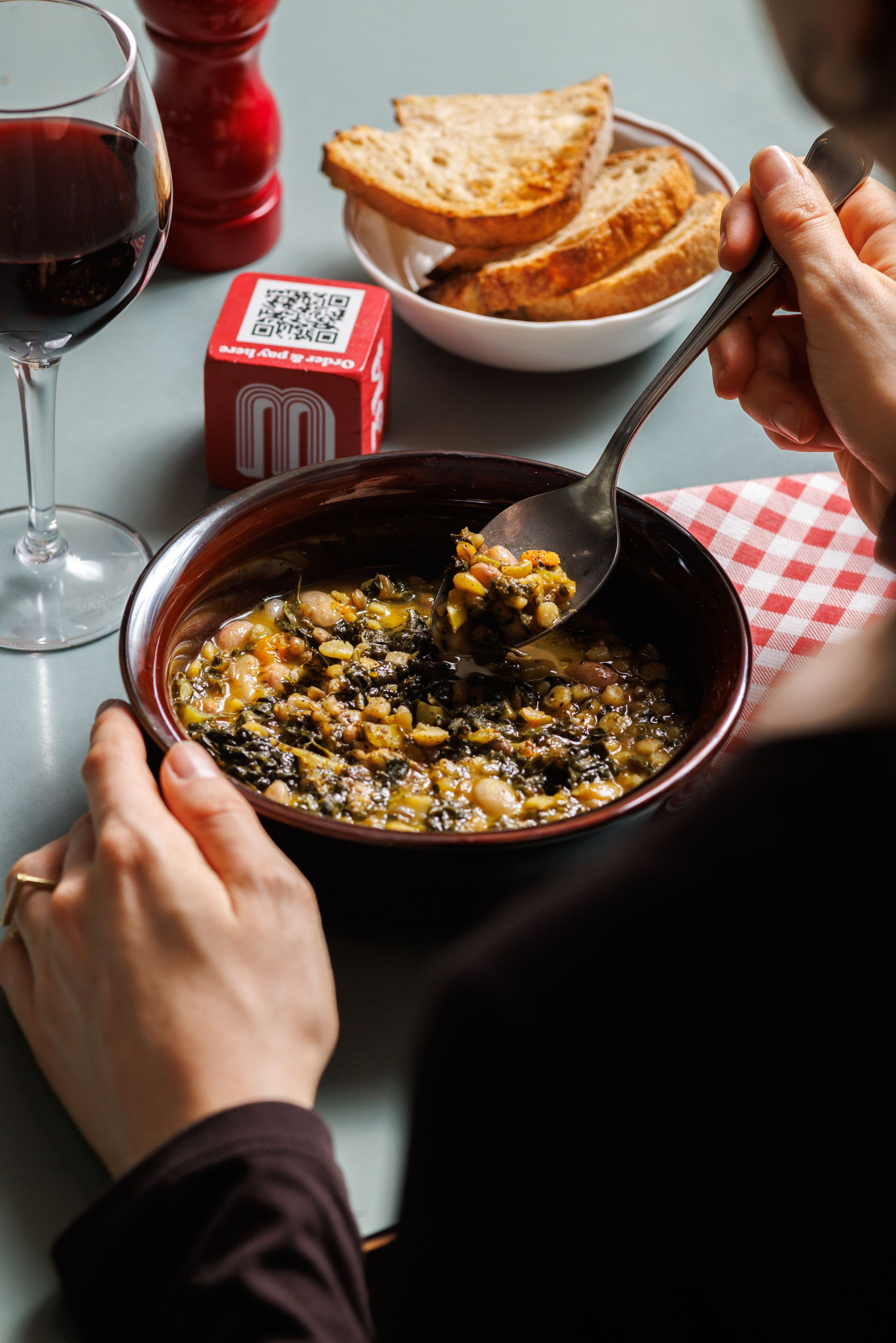 A person enjoys a hearty bowl of bean and kale soup accompanied by toasted bread and red wine in a cozy restaurant setting.