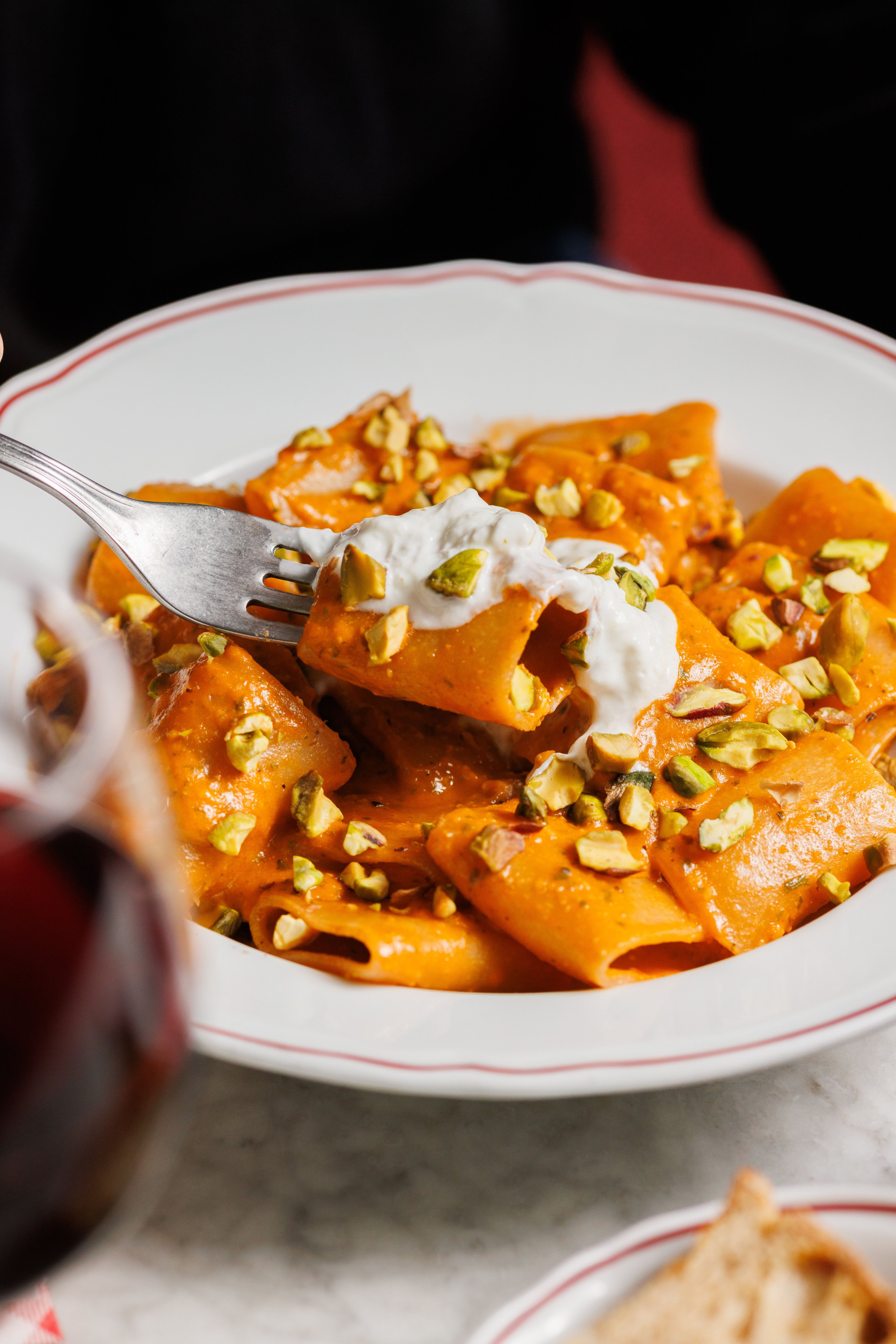 A close-up shot of a fork lifting a piece of rigatoni pasta coated in a creamy orange sauce and topped with crushed pistachios.