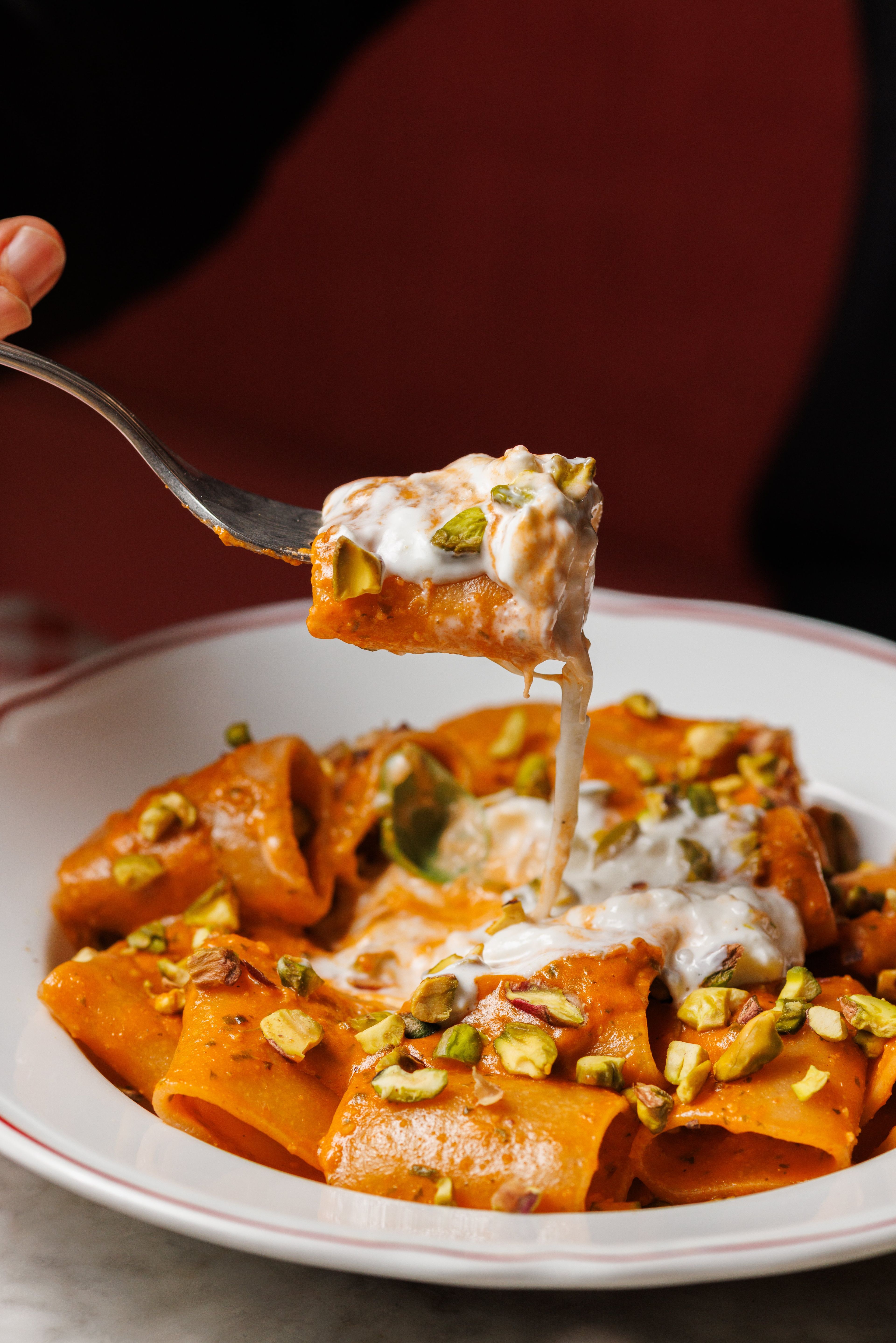 A close-up of a fork lifting a piece of rigatoni pasta coated in a creamy orange sauce and topped with crushed pistachios.