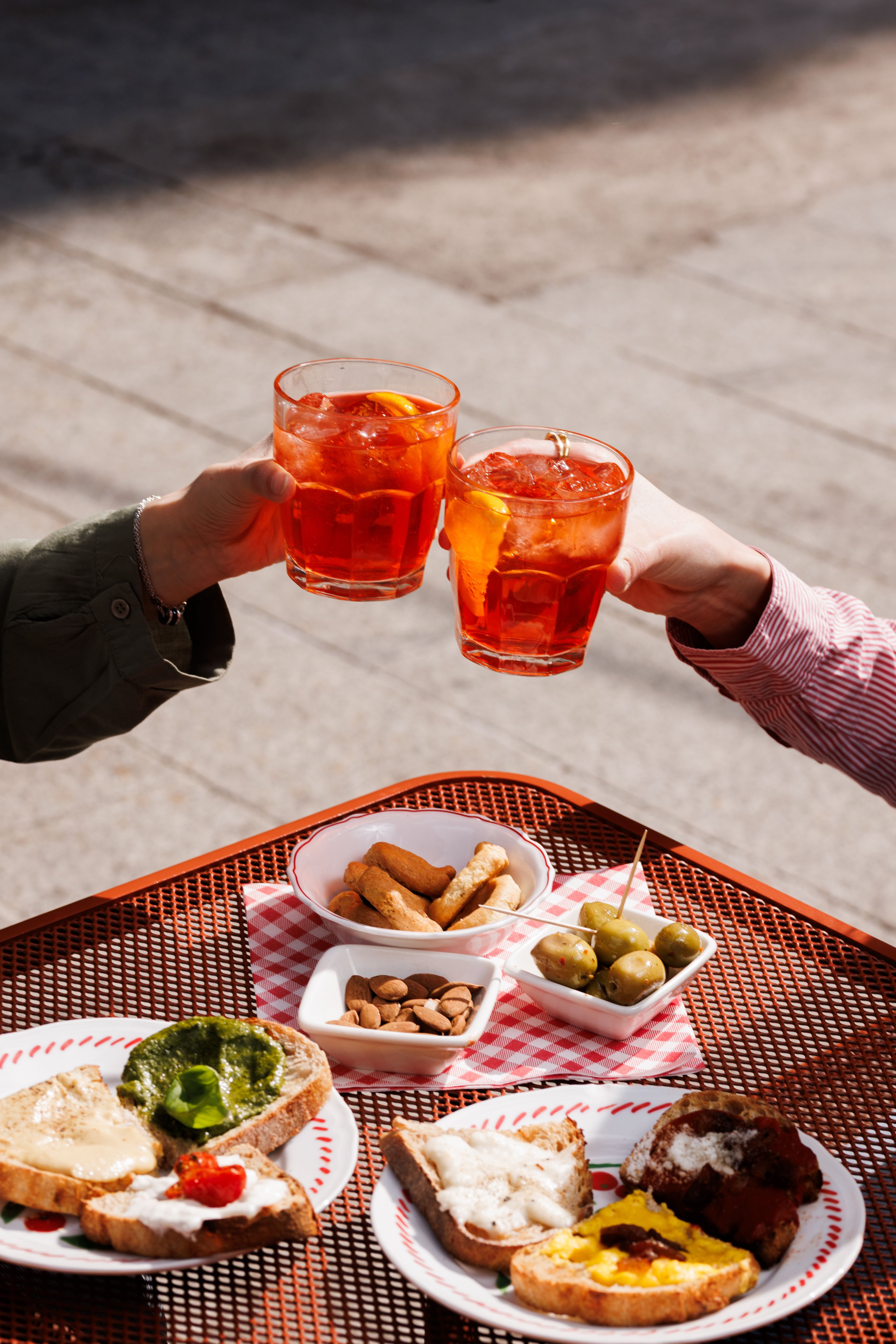 Two people toast with Aperol spritzes over a table filled with various bruschetta, olives, and almonds on a sunny patio.
