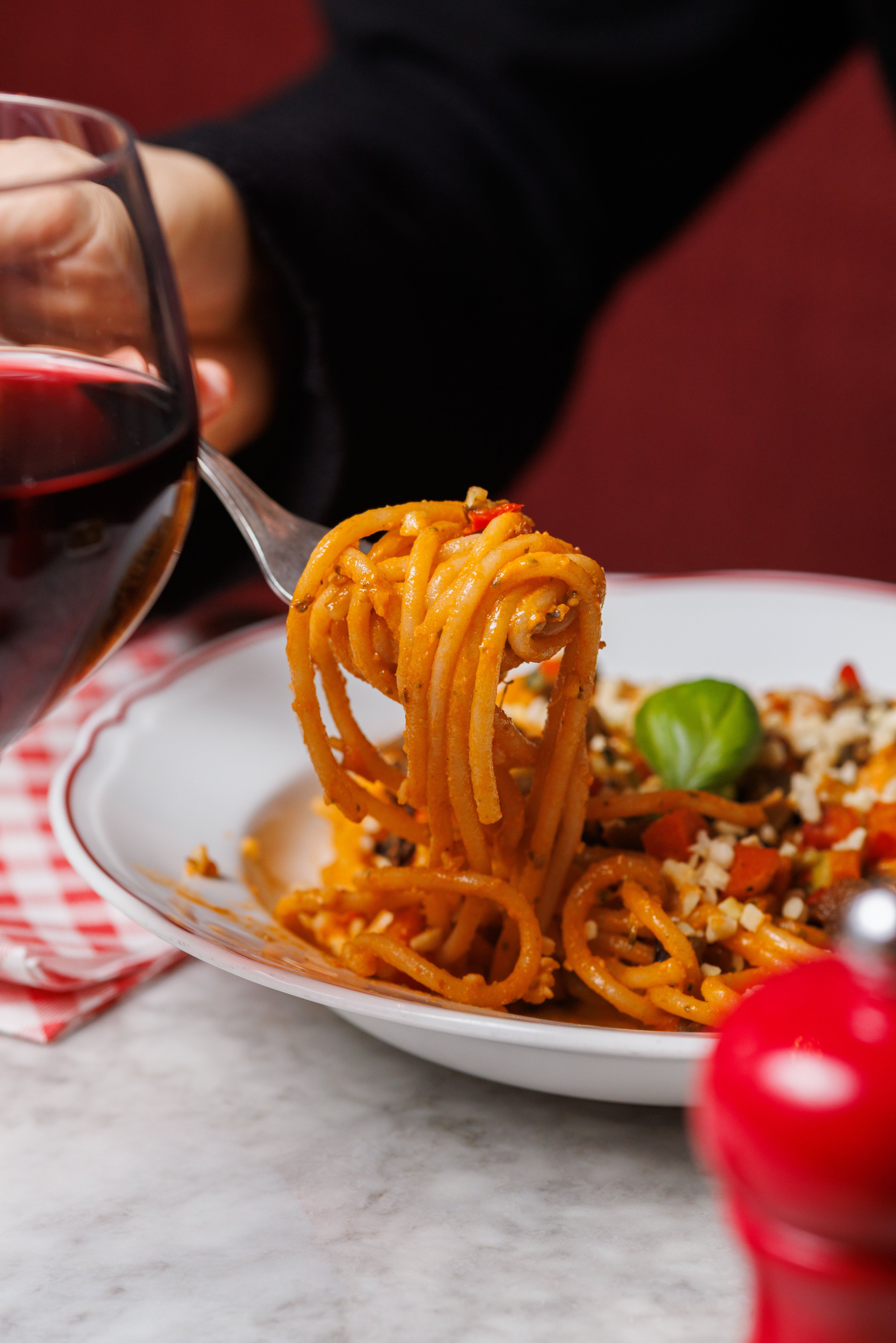 A close-up of a fork lifting spaghetti with tomato sauce and vegetables from a plate next to a glass of red wine.