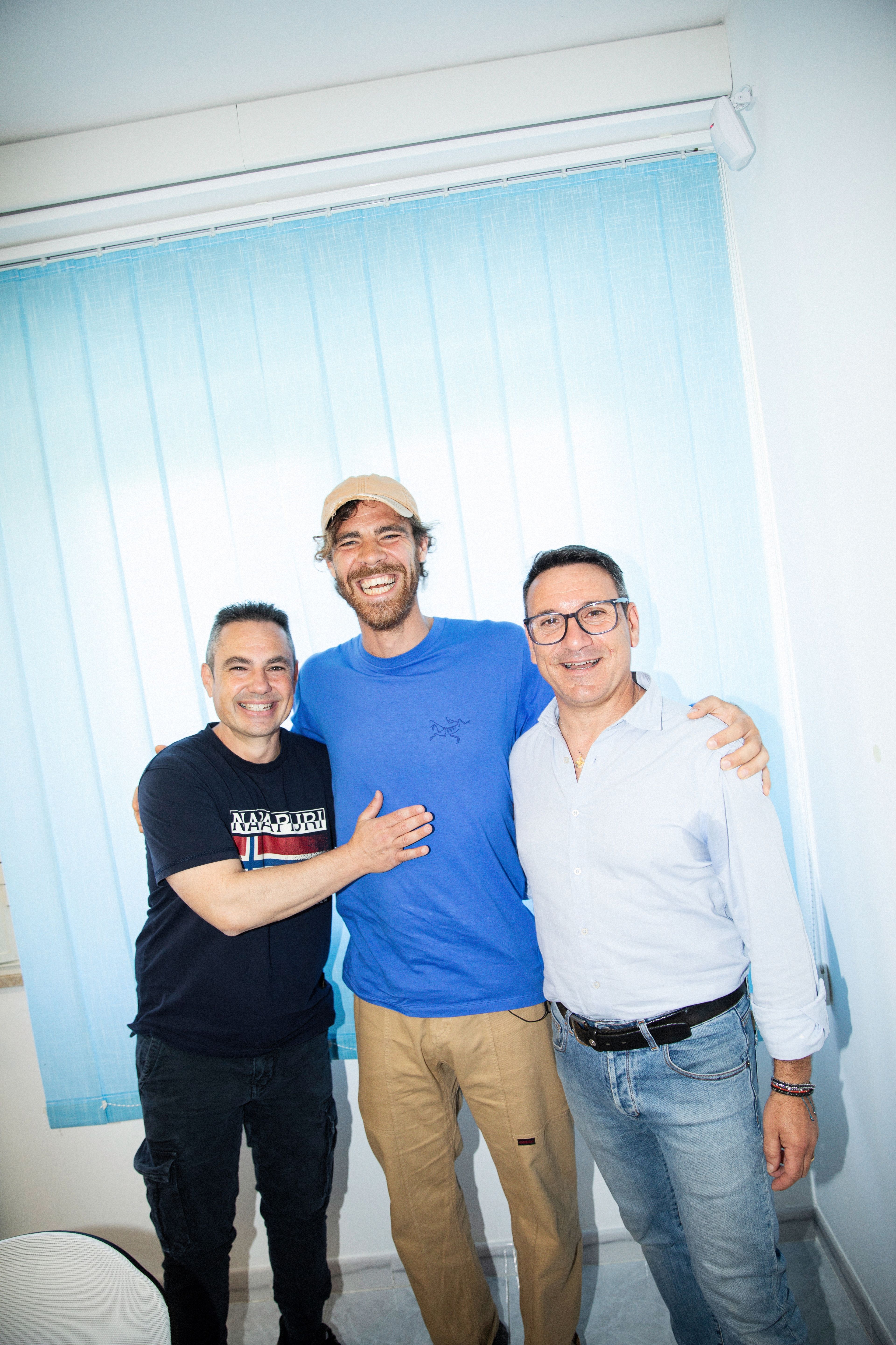 Three men are smiling and posing for a photo indoors against a light blue window blind.