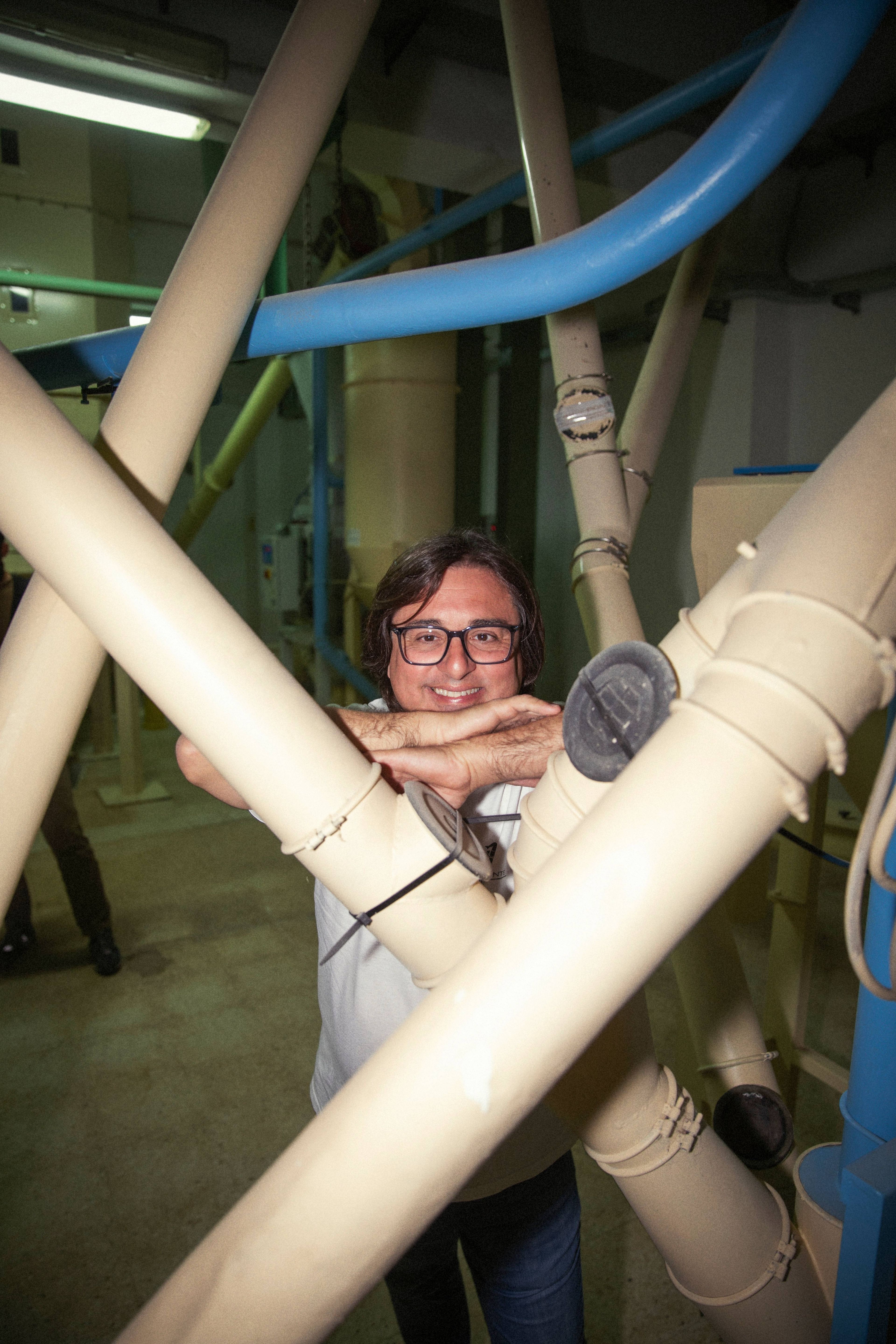 A man smiles while posing behind industrial pipes in a brightly lit, utilitarian facility.