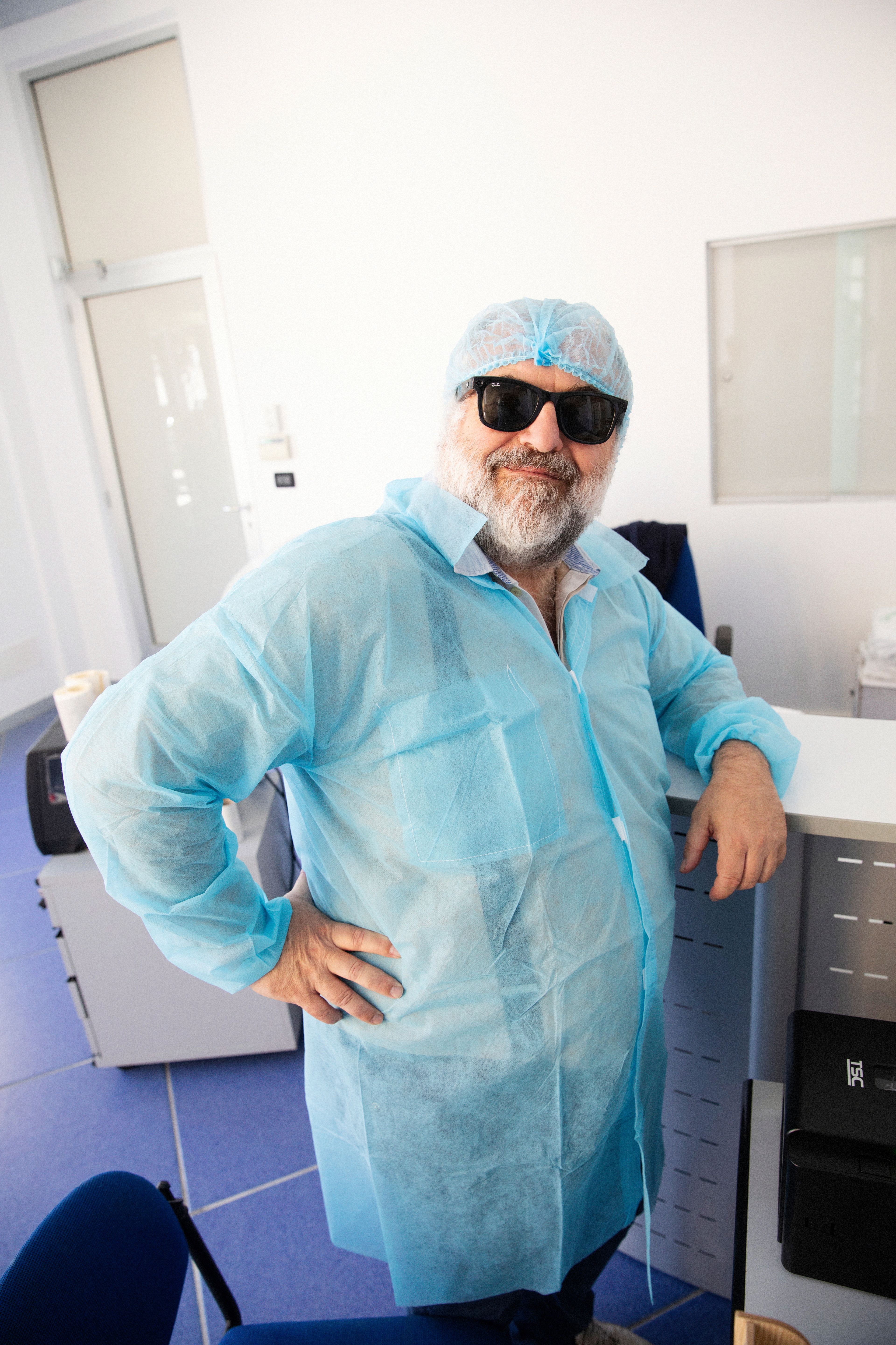 A man wearing a protective gown, hairnet, and sunglasses stands in a sterile, brightly lit office environment.