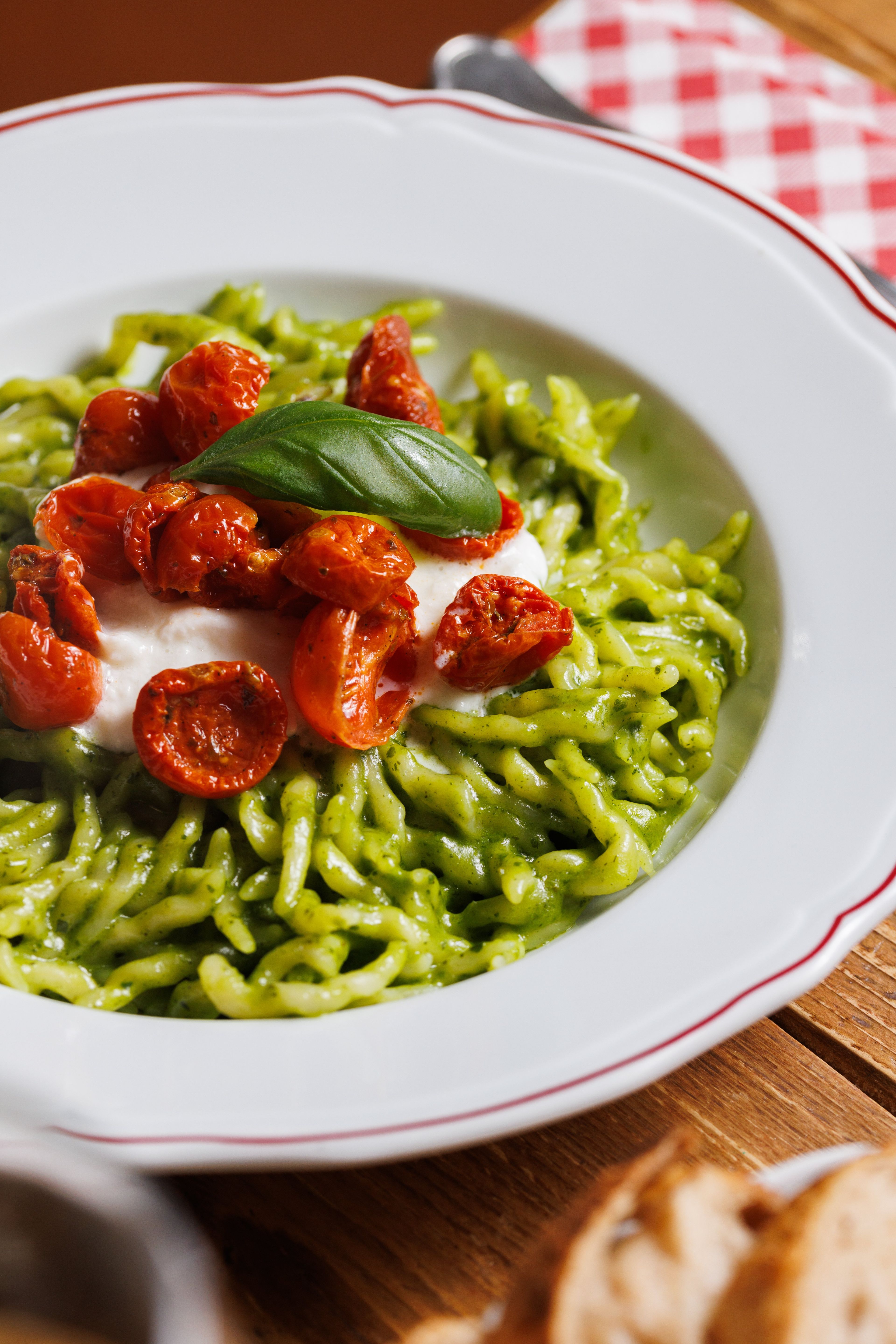 A plate of fresh pasta tossed in vibrant green pesto sauce and topped with roasted cherry tomatoes, creamy cheese, and a fresh basil leaf.