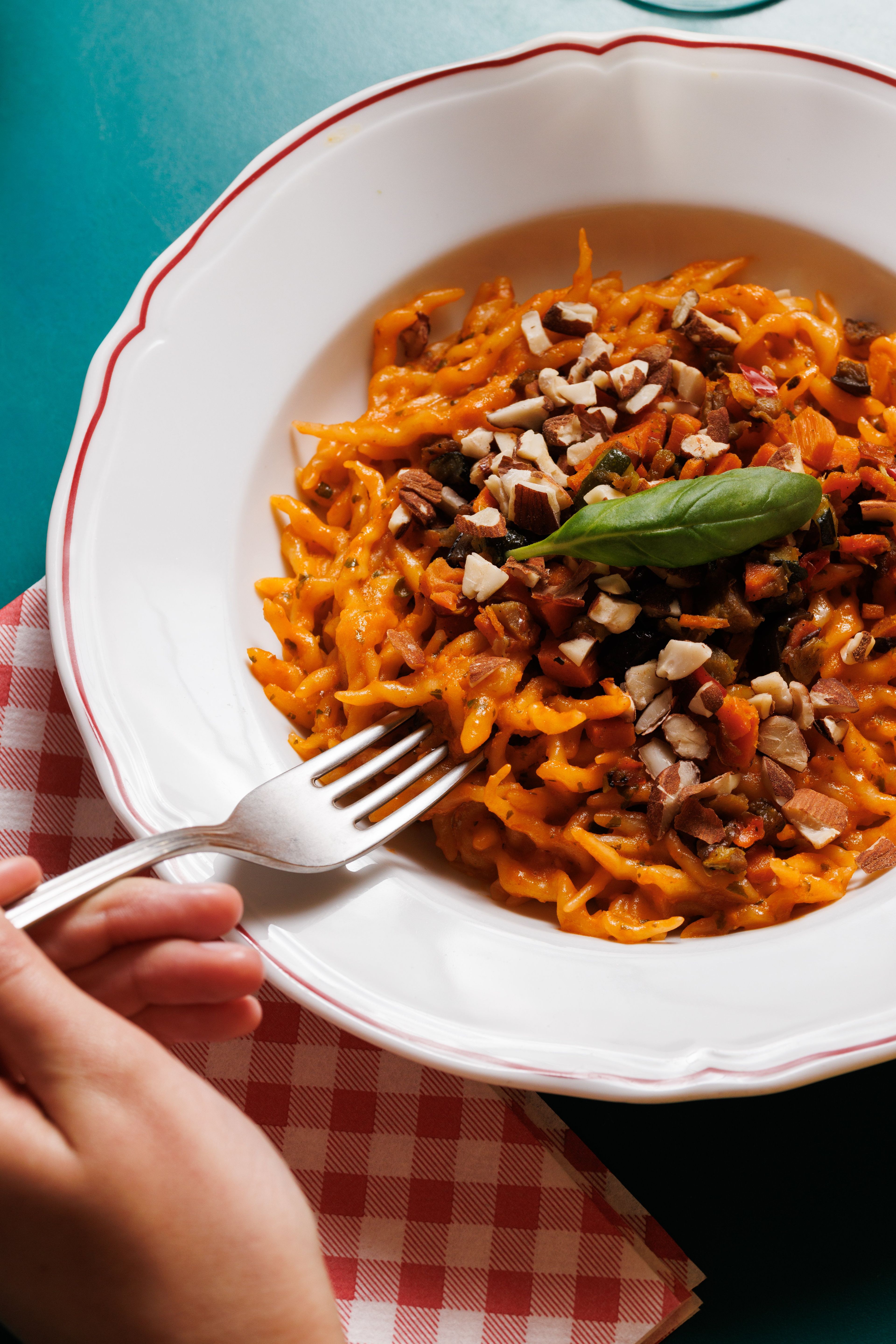 A hand holds a fork over a plate of pasta tossed in a vibrant orange sauce and topped with chopped nuts and a fresh basil leaf.