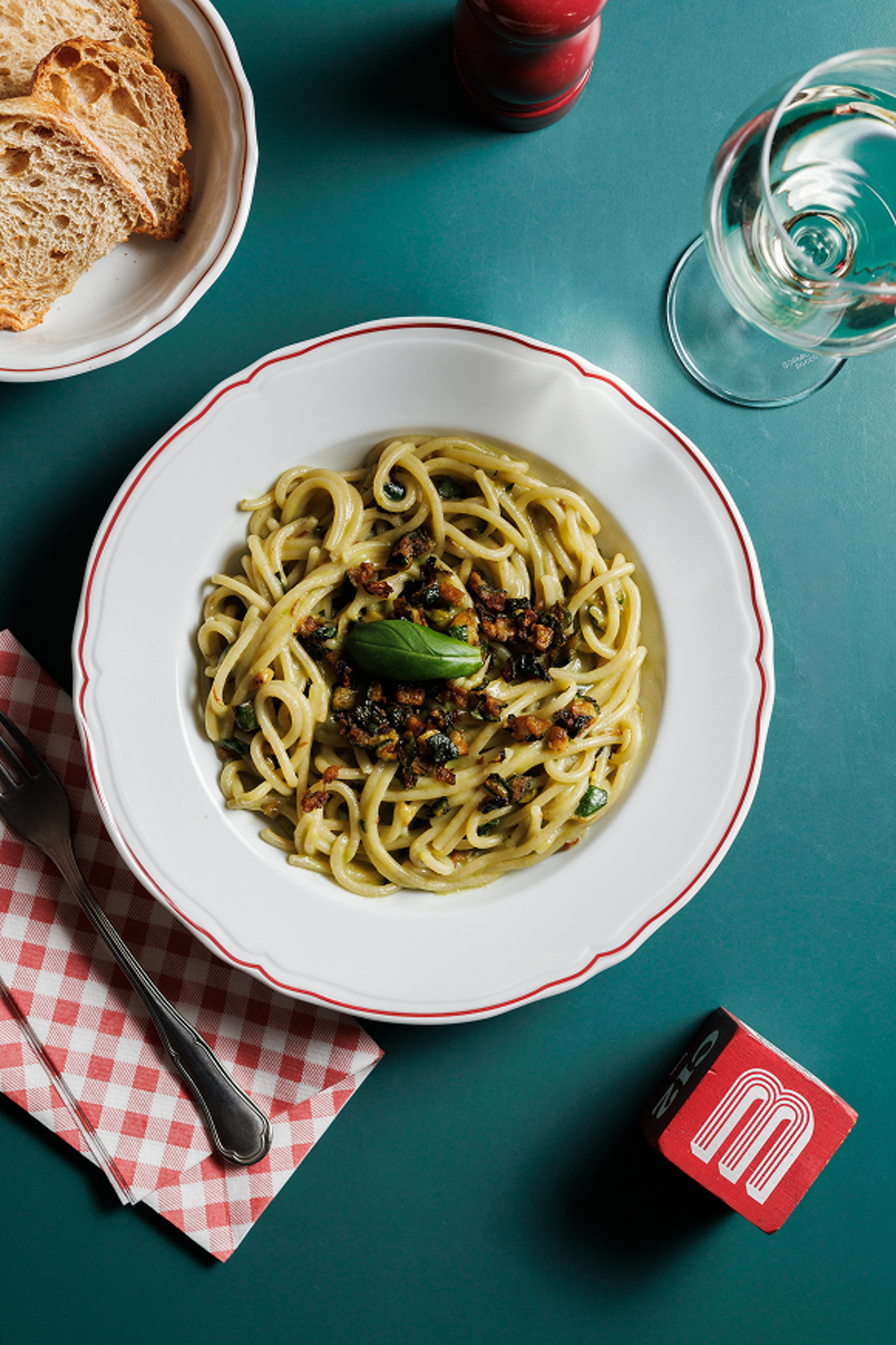 A top-down view of a plate of spaghetti with zucchini and basil served on a dark teal table with bread and wine.