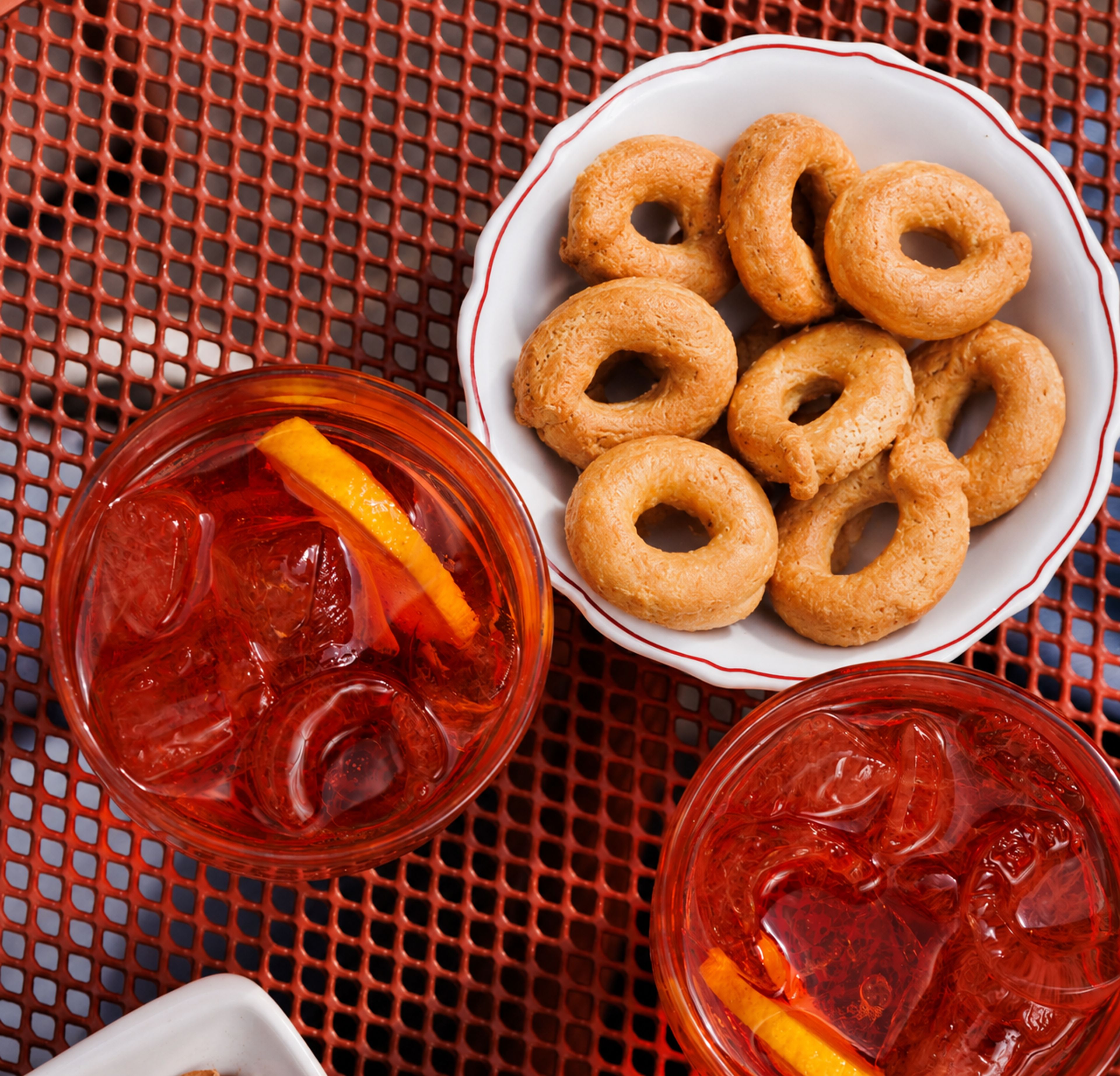 Two glasses of Aperol spritz are served alongside a small bowl of taralli crackers on a red metal table.
