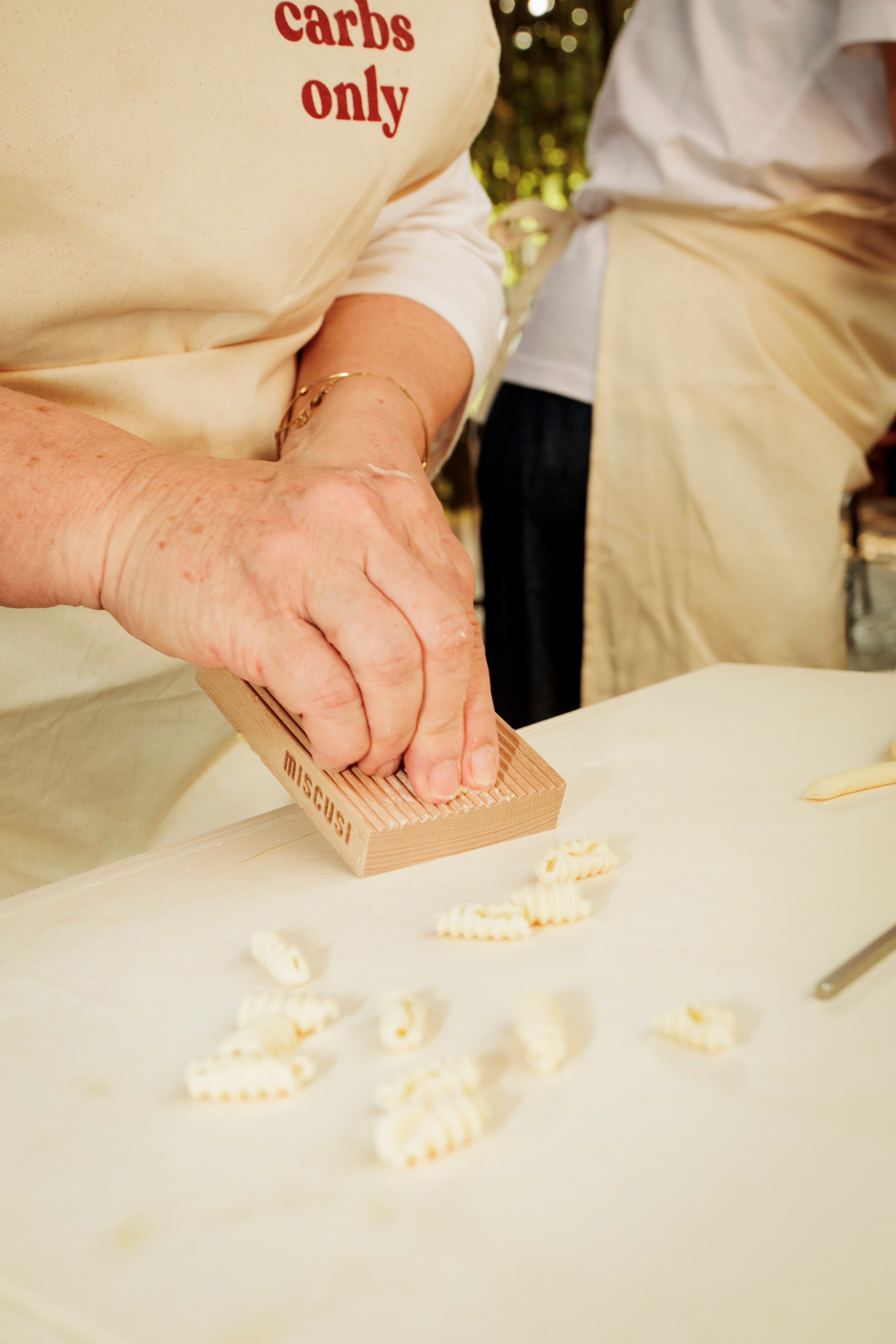 A person uses a wooden gnocchi board to shape fresh pasta dough on a white table.