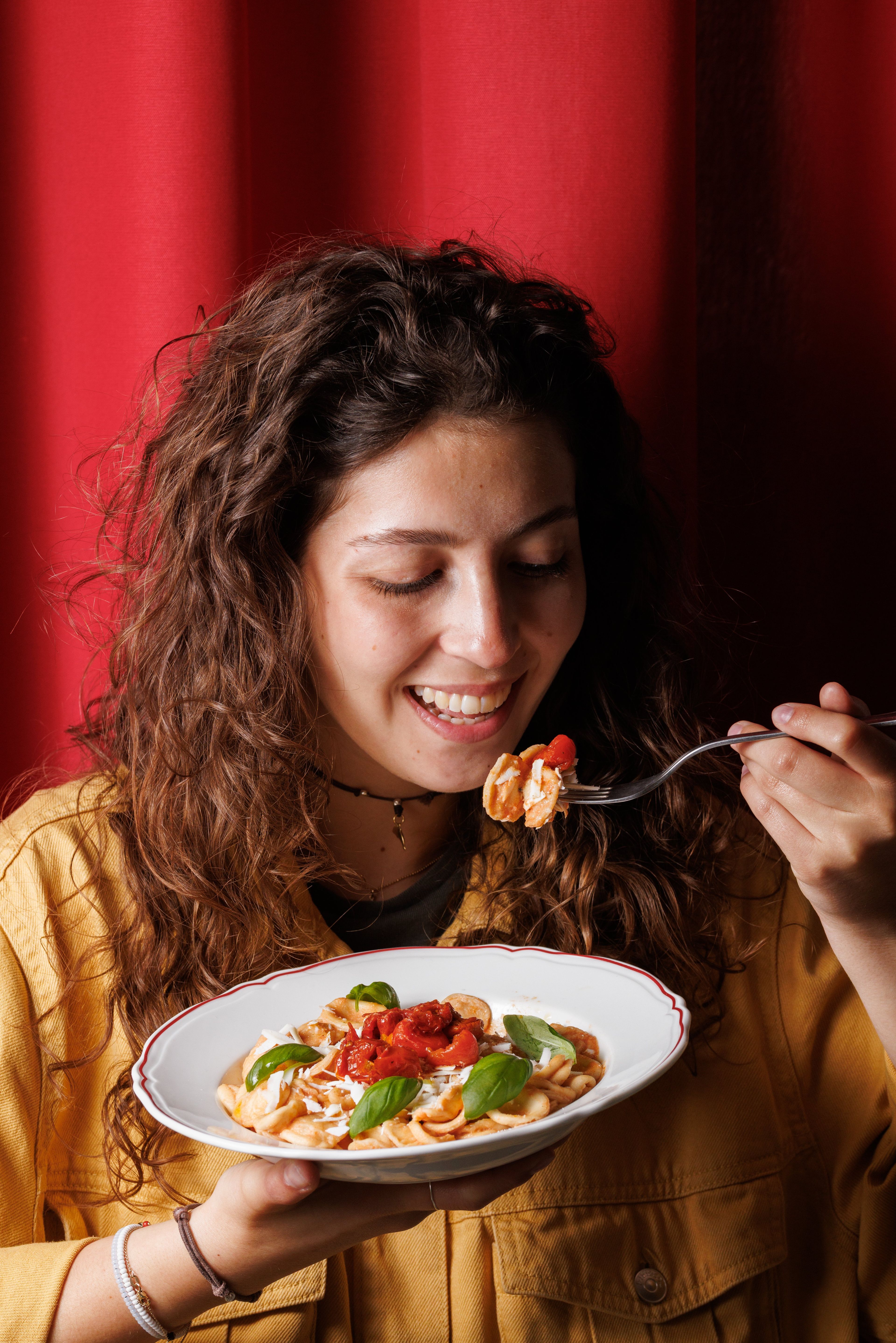 A smiling woman enjoys a plate of fresh orecchiette pasta topped with roasted tomatoes and basil in a warm, intimate setting.