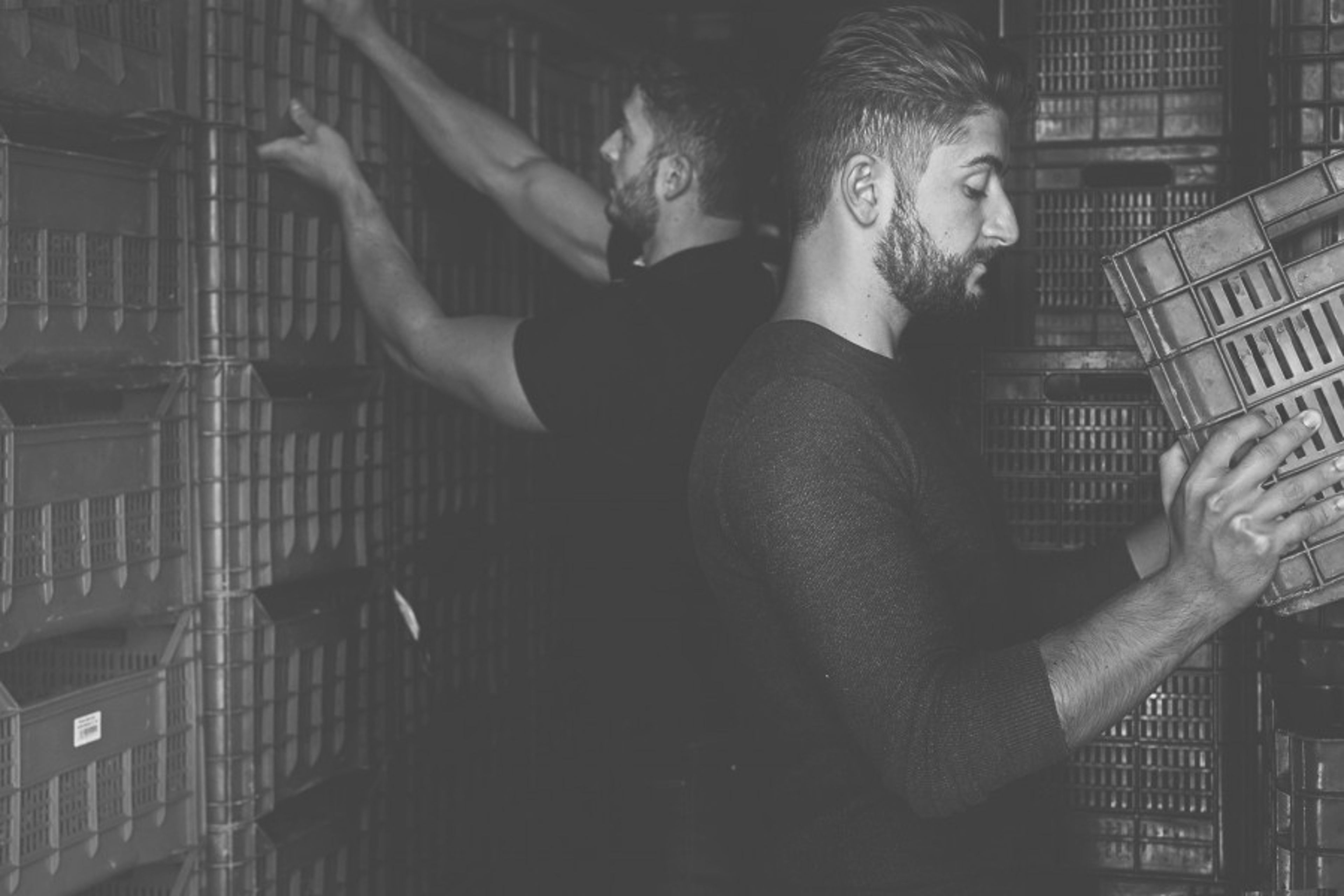 Two men work in a dimly lit storage area filled with stacked plastic crates.