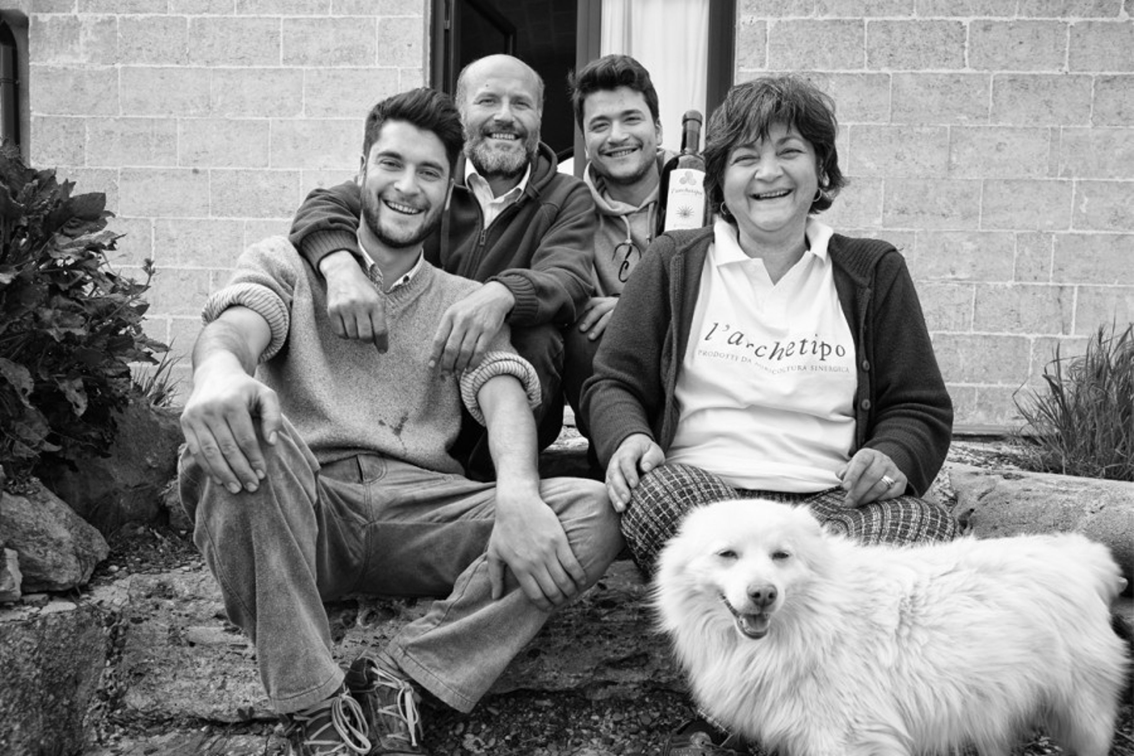 A family poses with their dog in front of a stone building while holding a bottle of wine.
