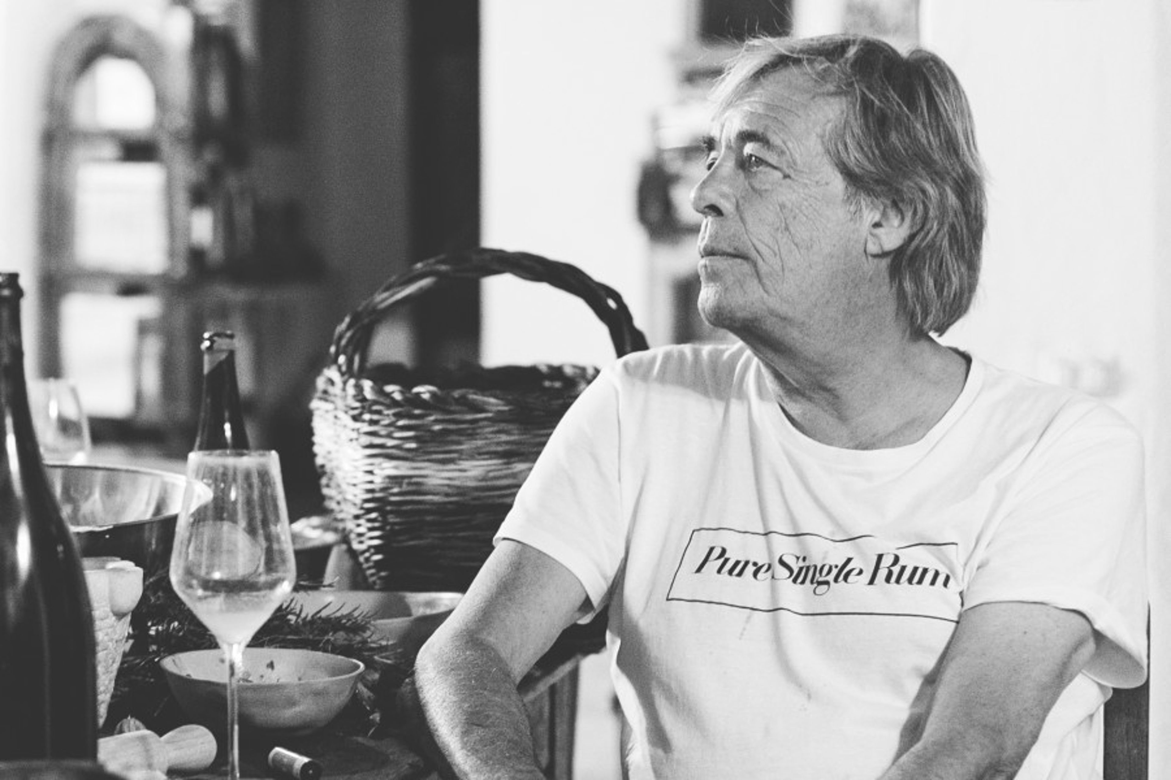A man wearing a t-shirt sits at a table with a wine glass and a basket in a dimly lit indoor setting.
