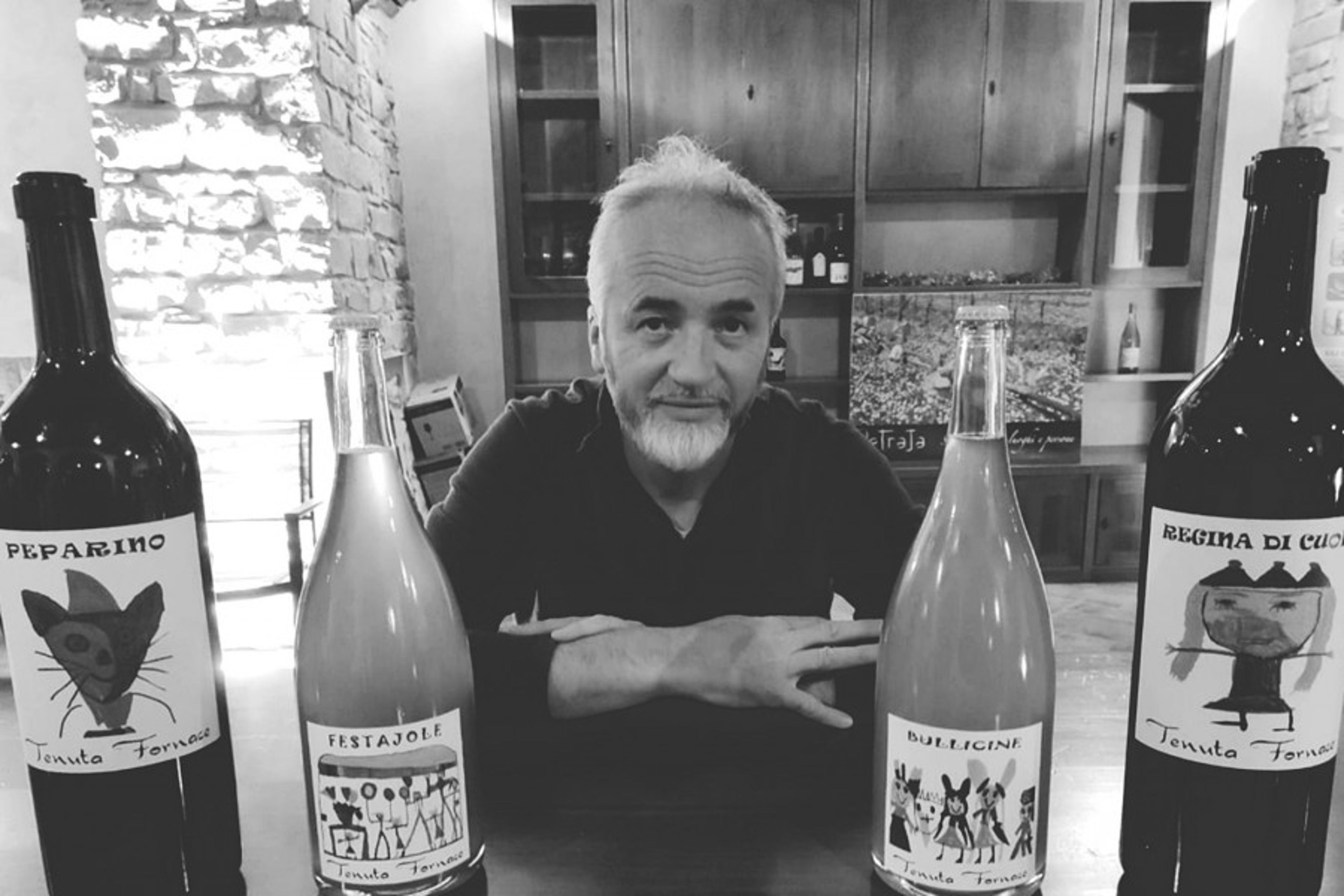 A man sits behind a table displaying four bottles of wine from Tenuta Fornace in a dimly lit, rustic setting.