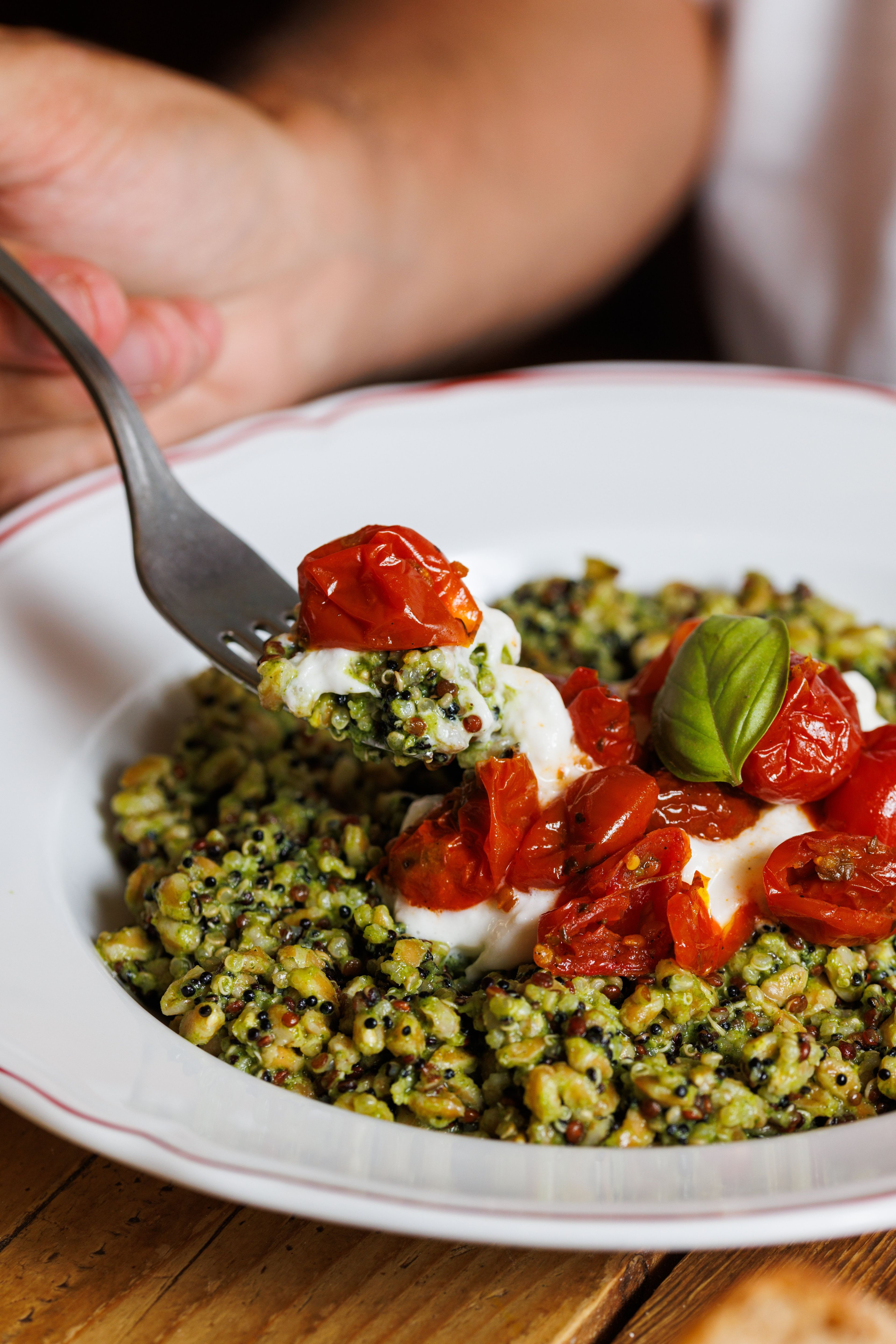 A close-up of a fork lifting a bite of pesto-coated quinoa salad topped with roasted cherry tomatoes and creamy cheese.