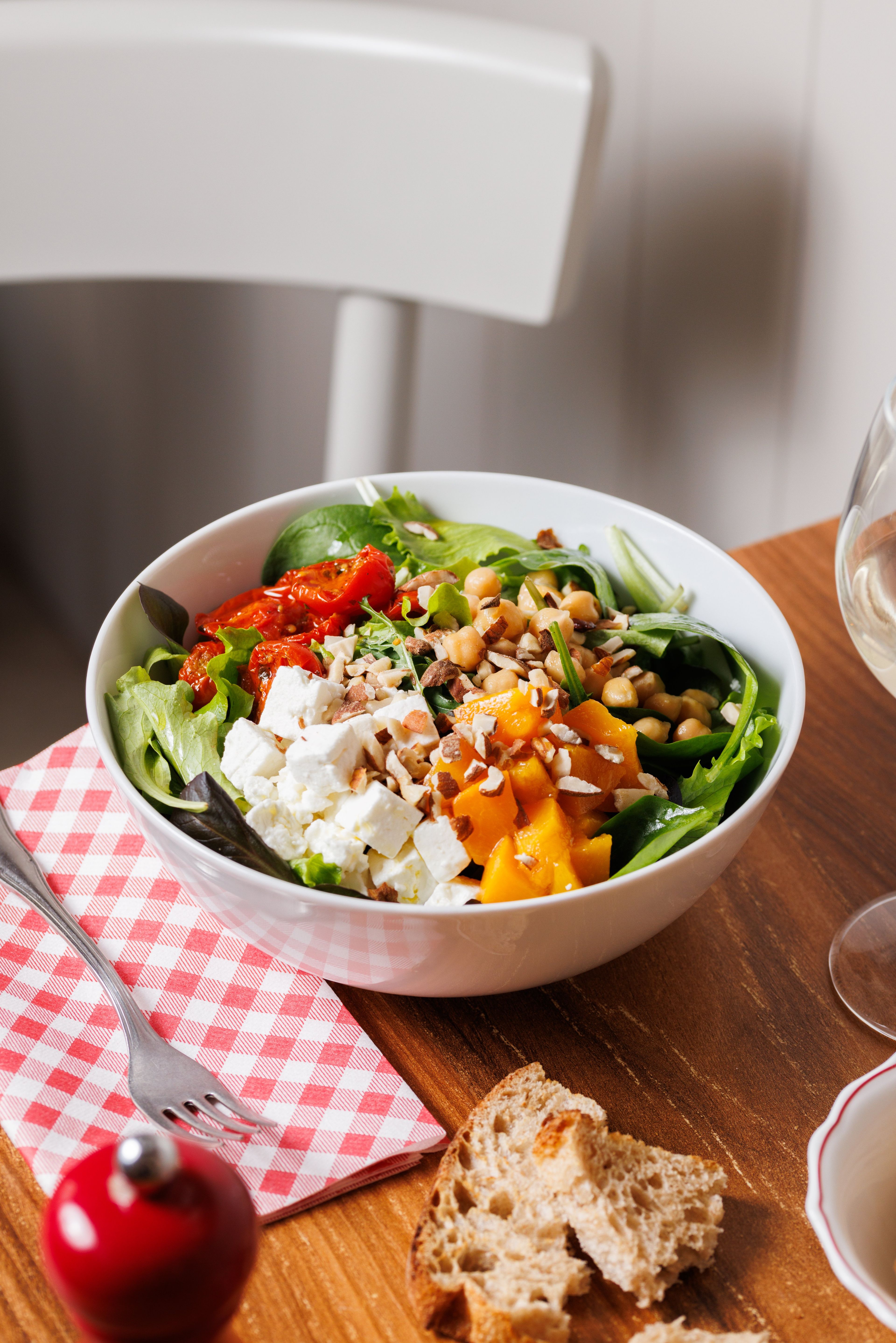 A fresh salad bowl featuring feta, chickpeas, roasted squash, and sun-dried tomatoes sits on a rustic wooden table.