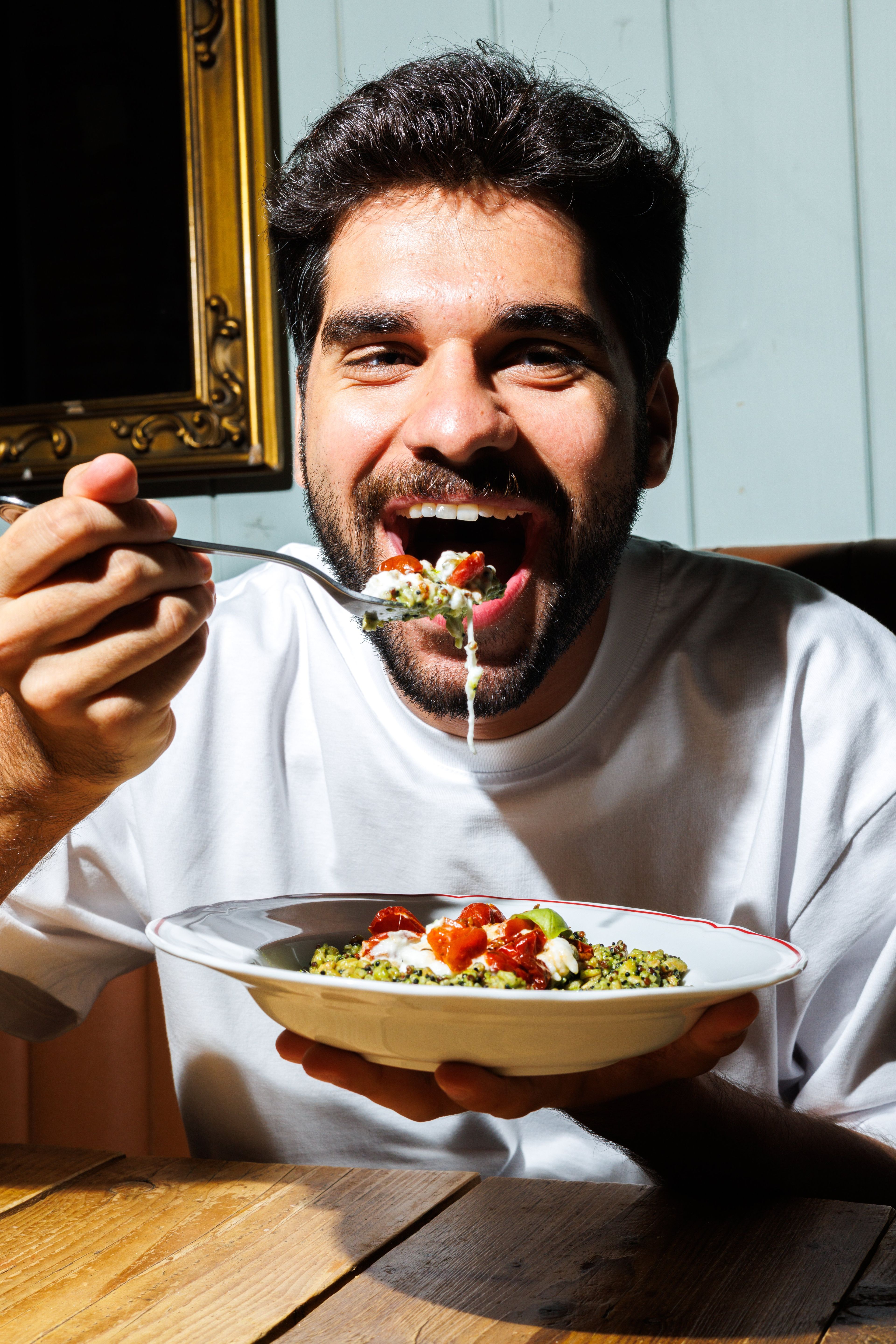 A man enjoys a vibrant bowl of pesto quinoa topped with sun-dried tomatoes and creamy cheese in a sunlit restaurant.