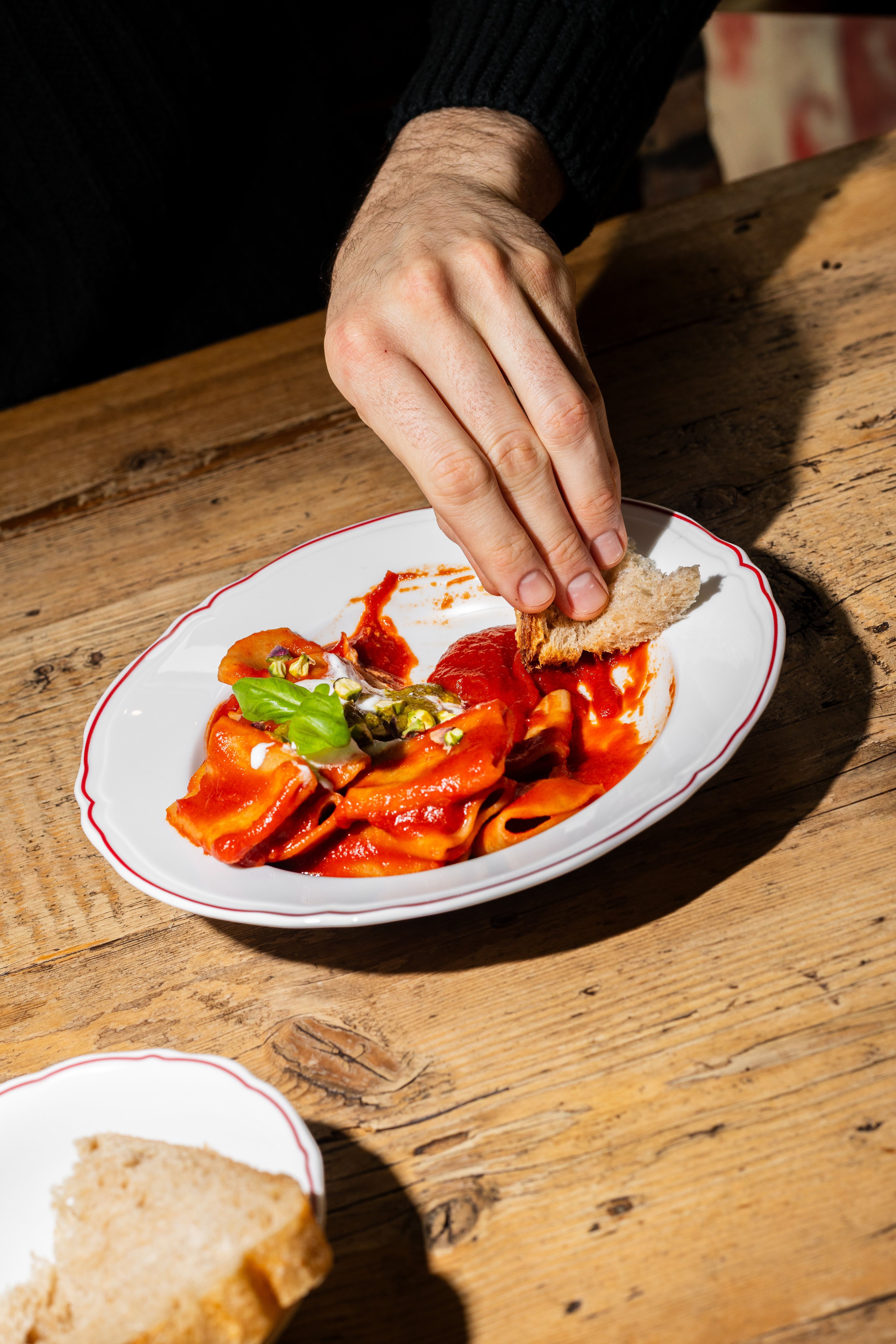 A person uses a piece of crusty bread to scoop up tomato-based pasta from a white plate on a rustic wooden table.