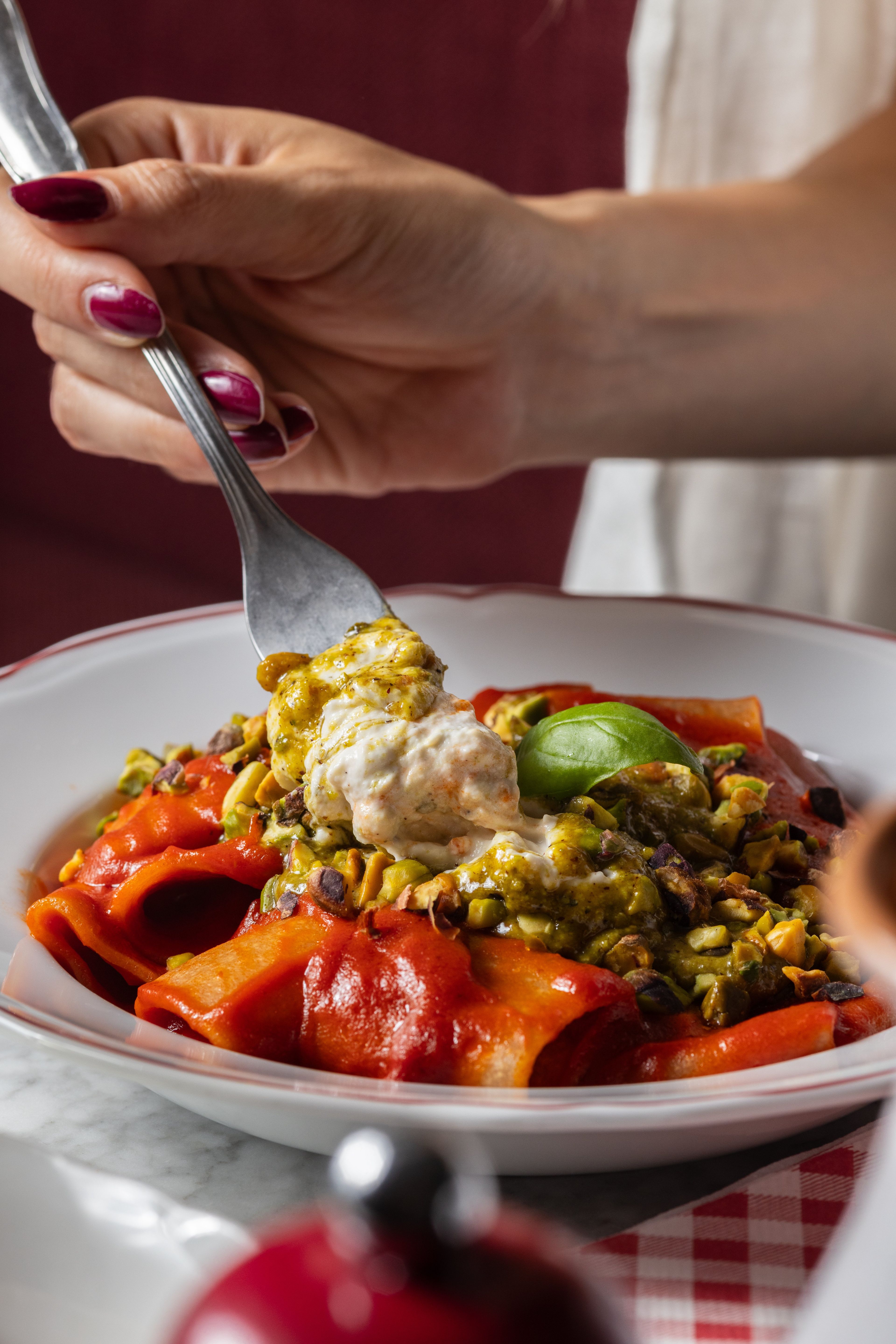 A hand uses a fork to lift a creamy, pistachio-topped pasta dish from a vibrant red sauce in a restaurant setting.