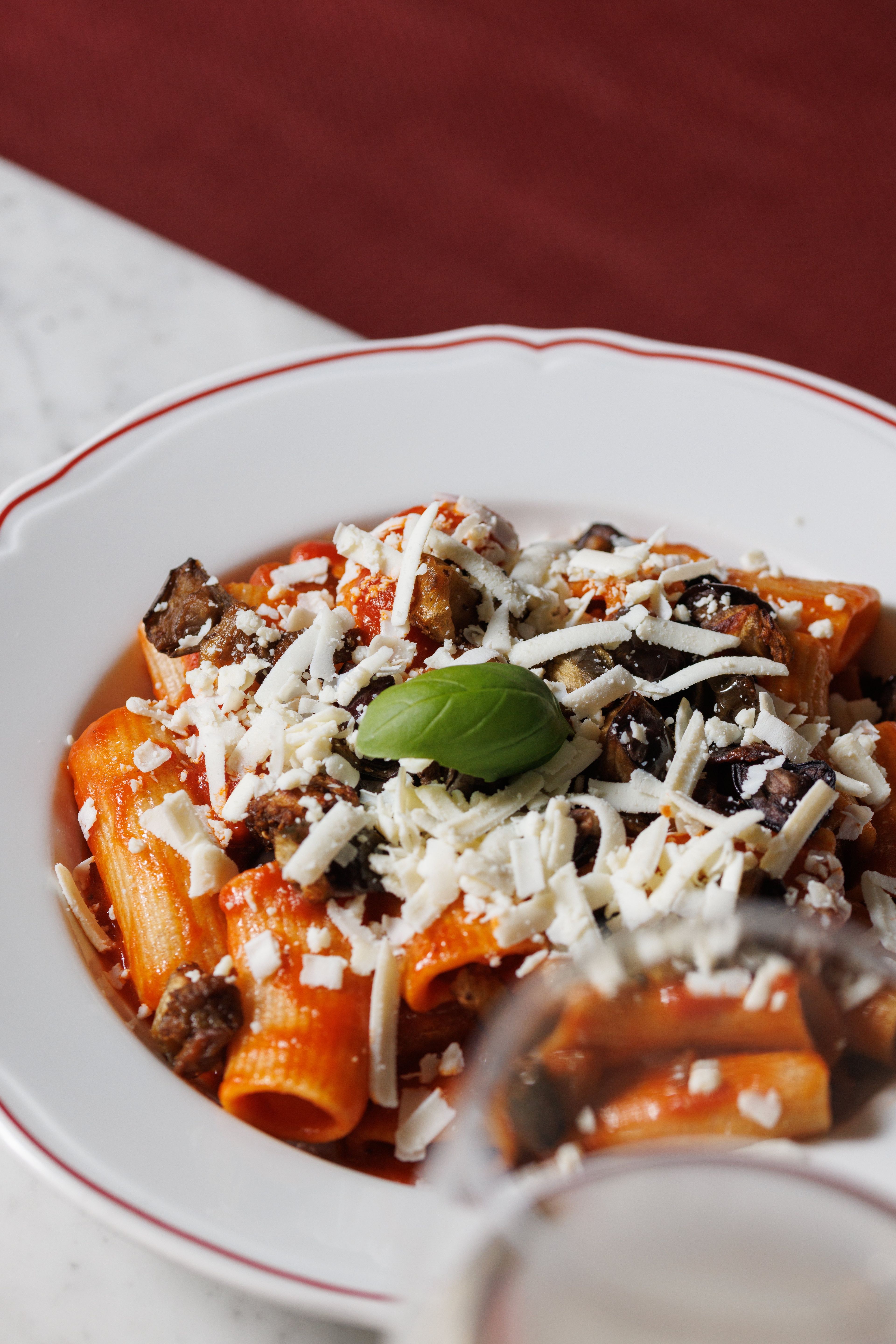 A plate of rigatoni pasta topped with tomato sauce, roasted eggplant, grated cheese, and a fresh basil leaf.