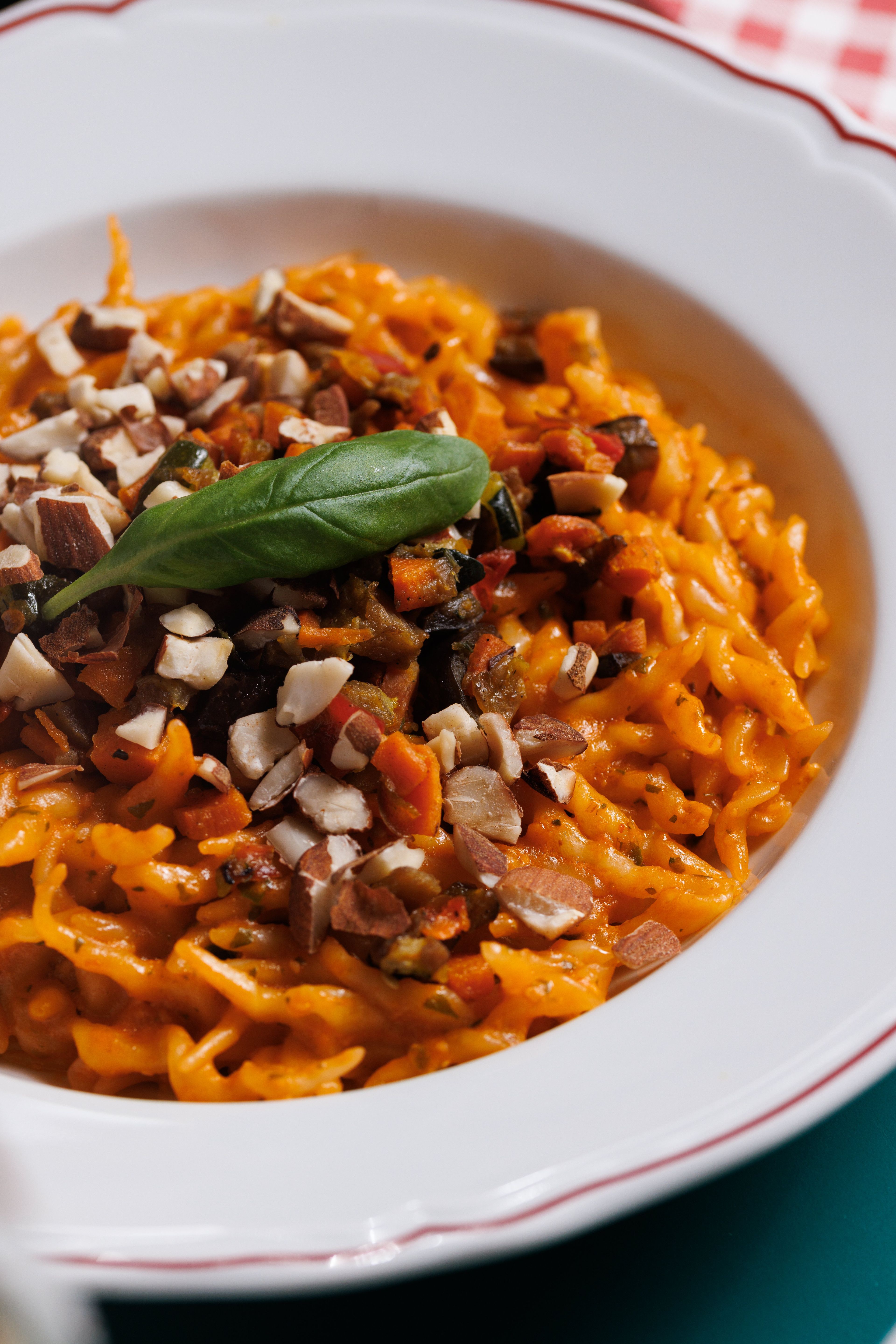 A close-up of a bowl of pasta tossed in a vibrant orange sauce and topped with chopped nuts and a fresh basil leaf.