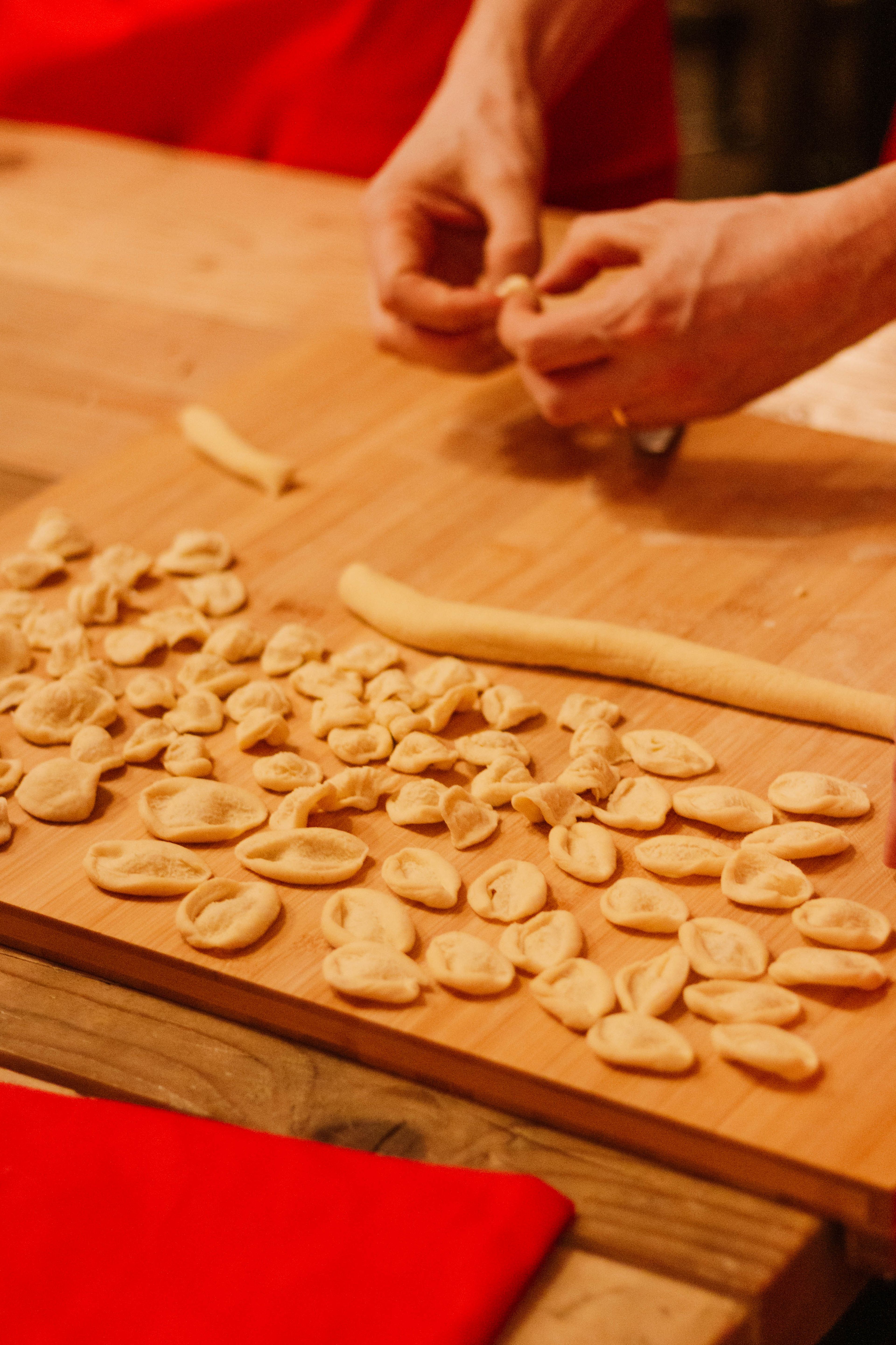 A person hand-shapes fresh orecchiette pasta on a wooden board in a warm, dimly lit kitchen.