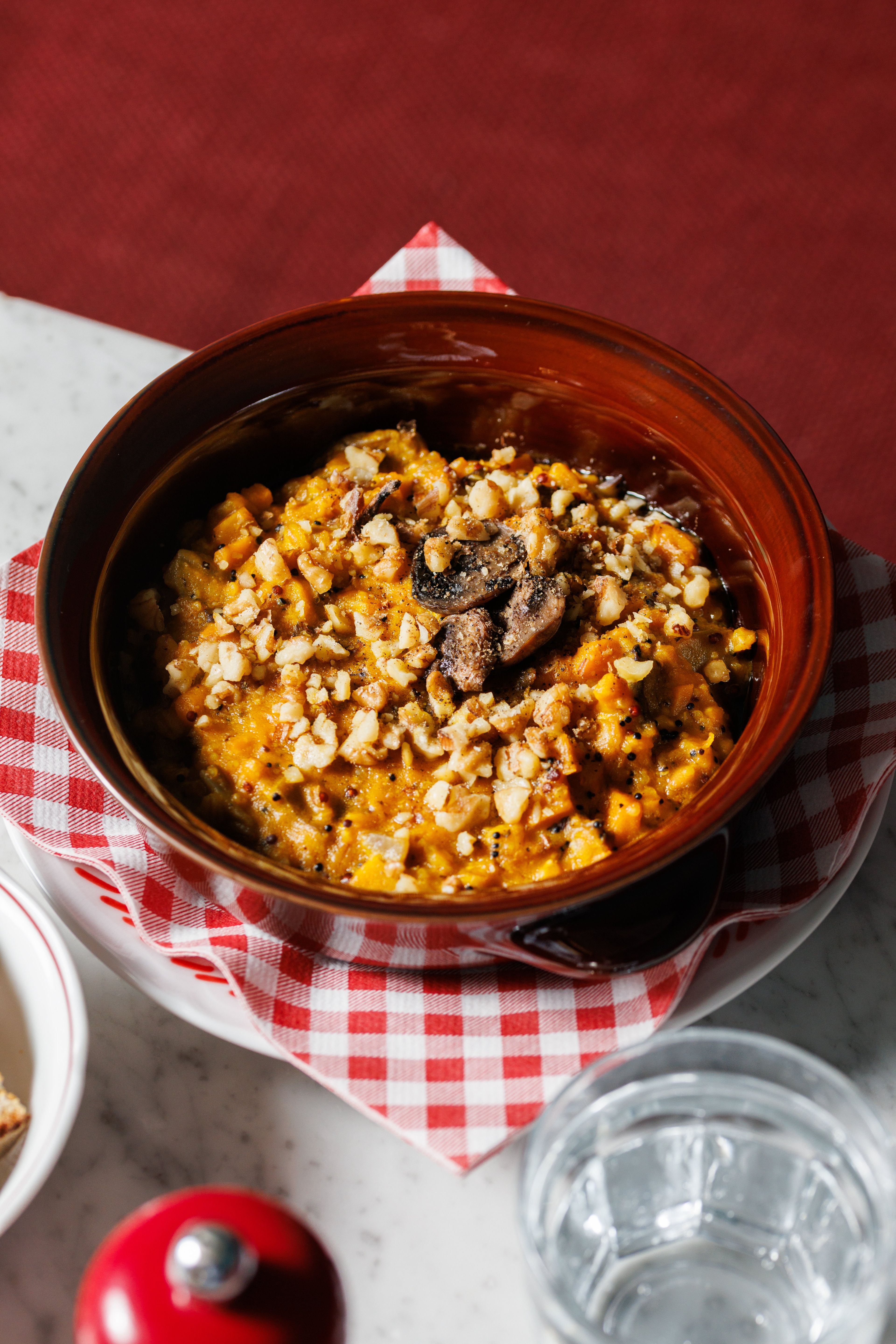 A rustic bowl of roasted pumpkin and mushroom risotto topped with chopped walnuts is served on a checkered napkin.