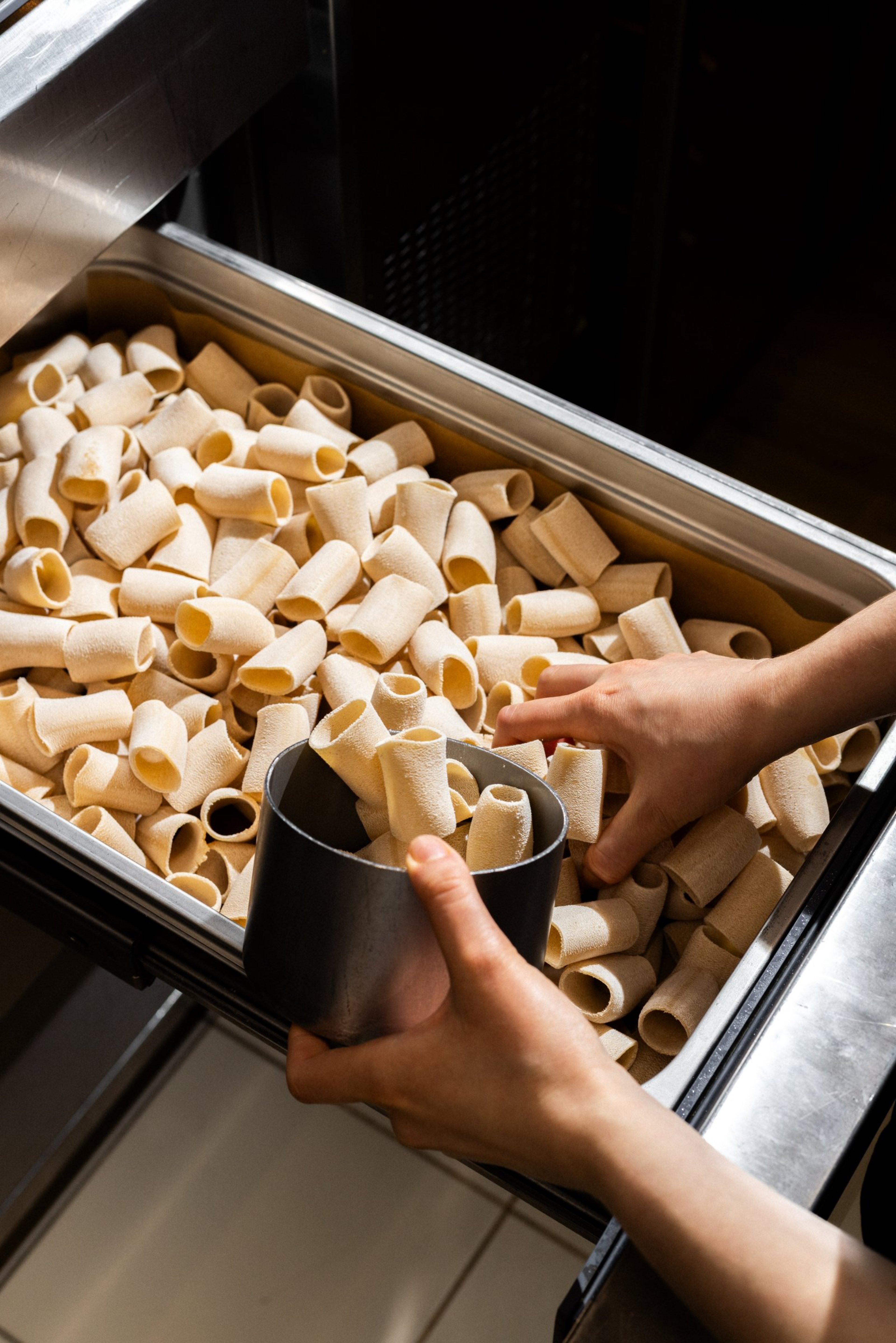 A chef uses a metal cup to scoop uncooked rigatoni pasta from a large stainless steel container.