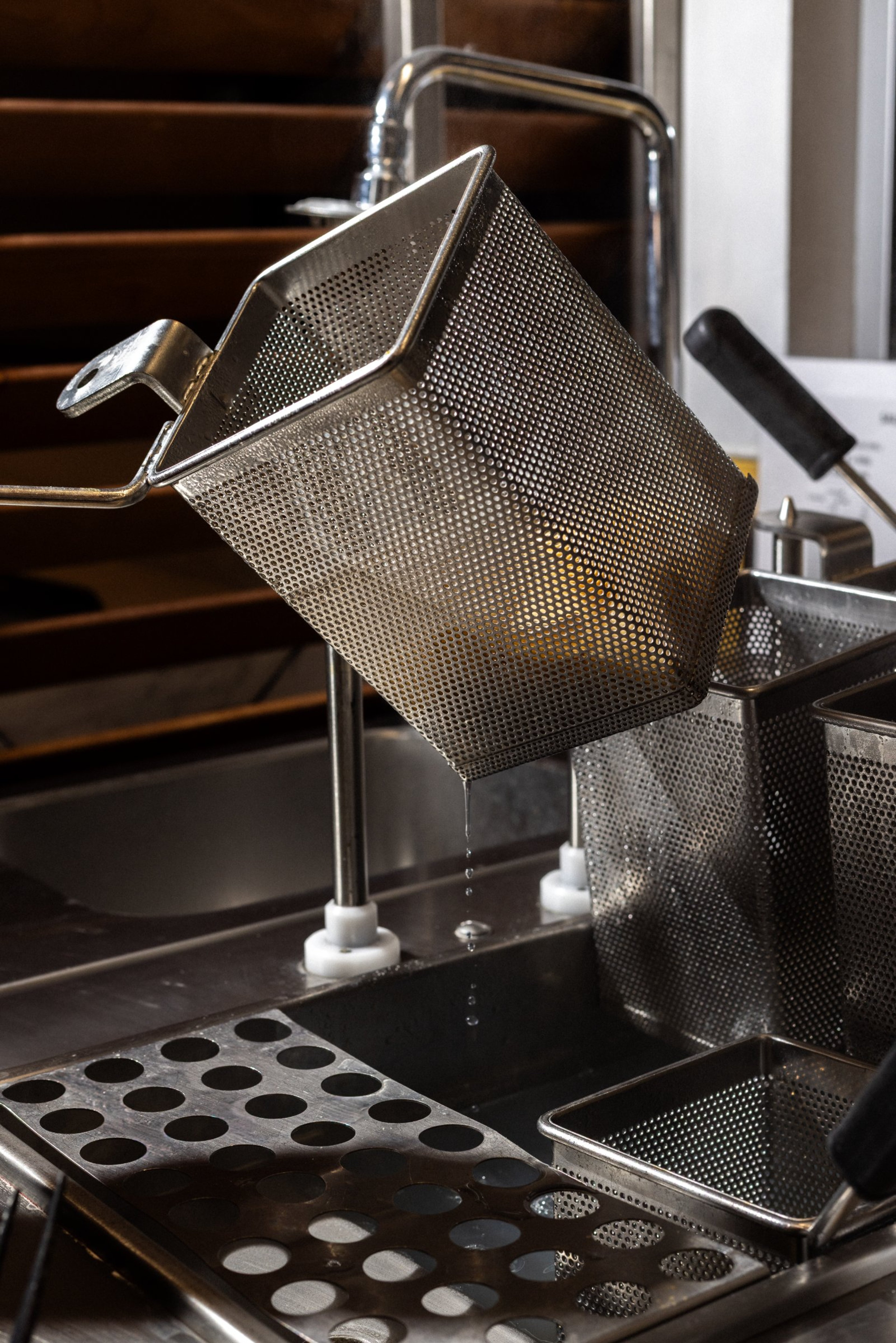 A metal fryer basket hangs over a commercial kitchen sink while dripping water.