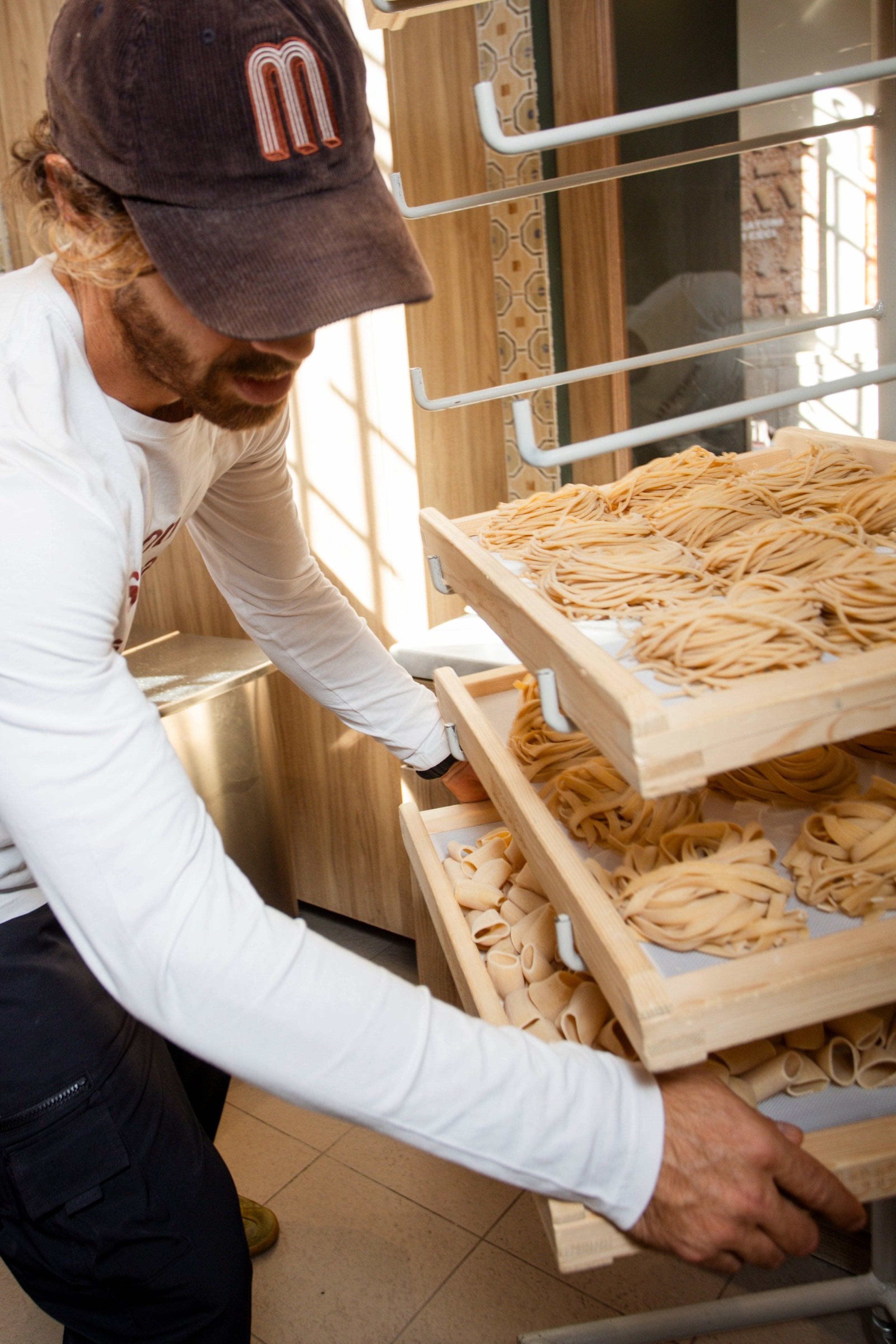 A chef carefully handles wooden trays filled with freshly made artisanal pasta in a brightly lit kitchen.