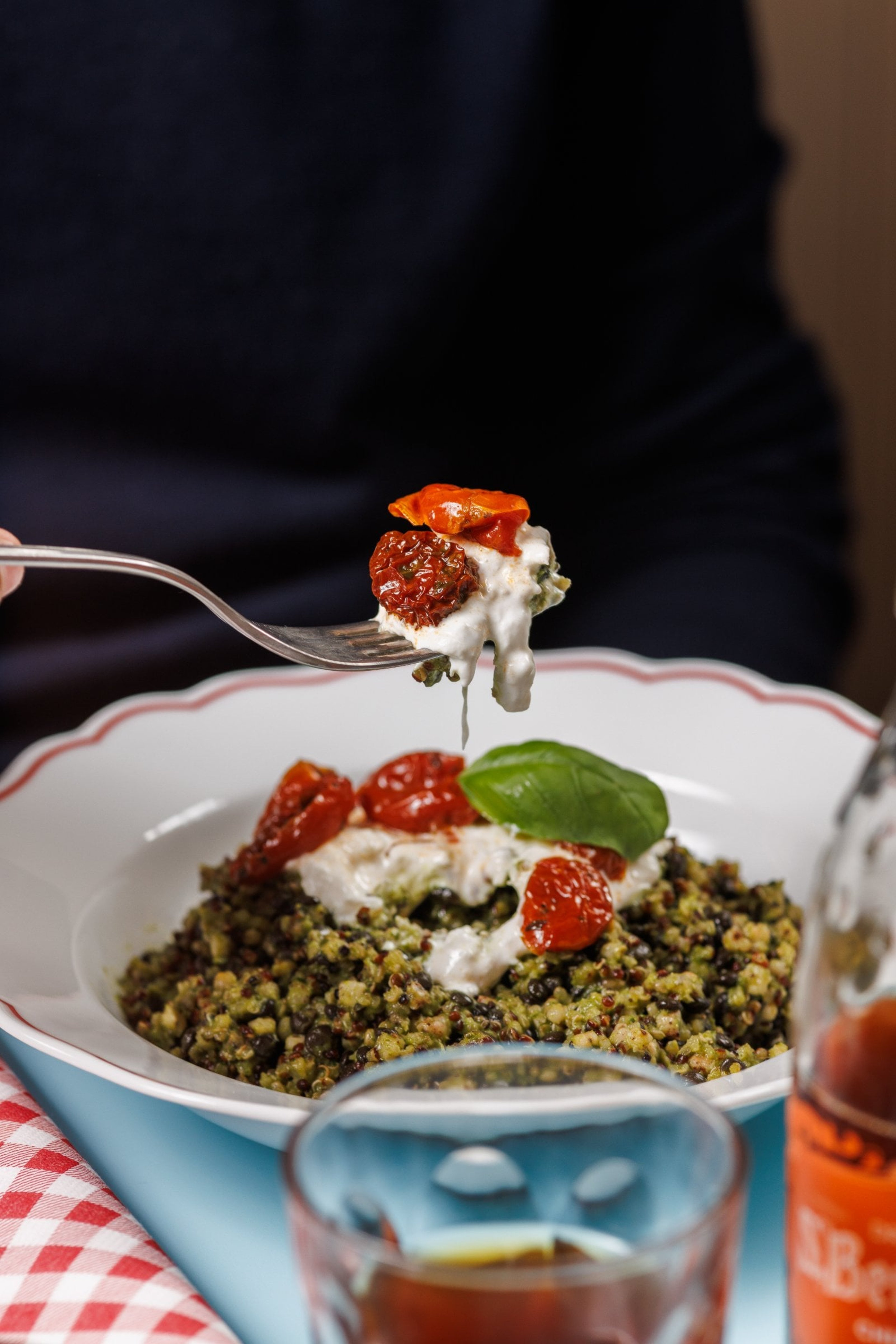 A fork lifts a bite of pesto-tossed quinoa topped with creamy burrata and sun-dried tomatoes from a white plate.