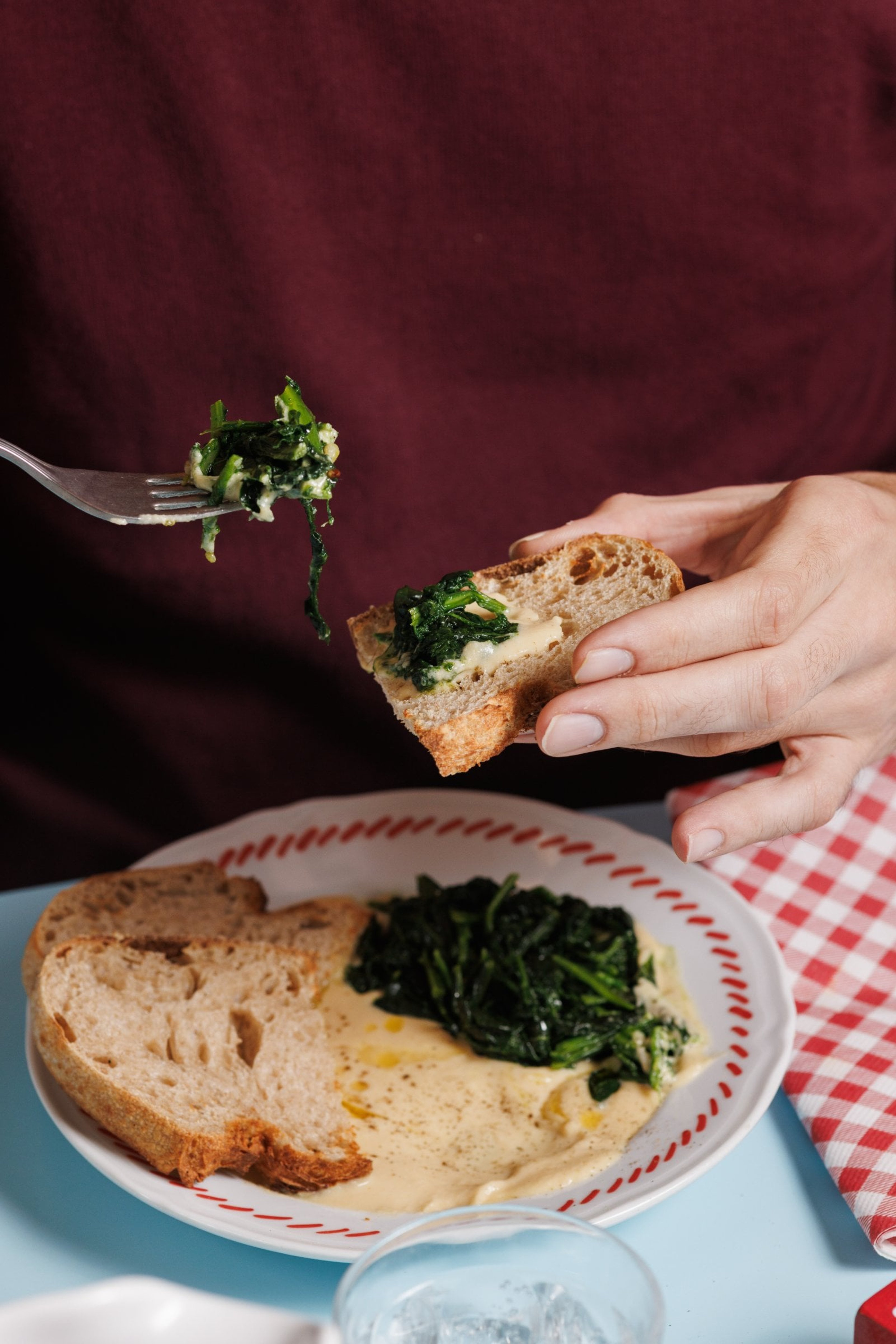 A person holds a piece of crusty bread topped with sautéed spinach and creamy sauce above a plate of the same meal.