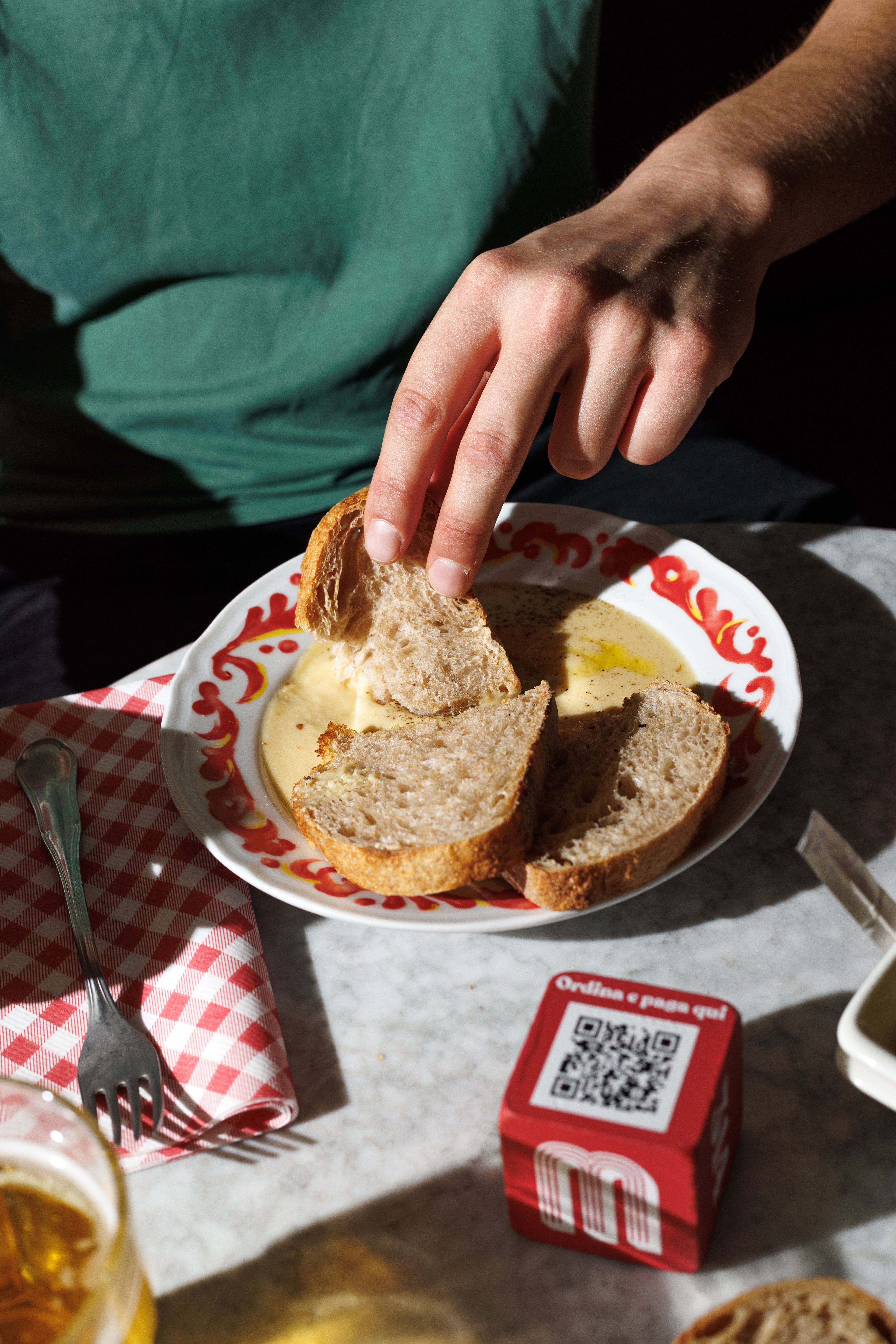 A person dips a slice of rustic bread into a bowl of creamy soup on a sunny restaurant table.
