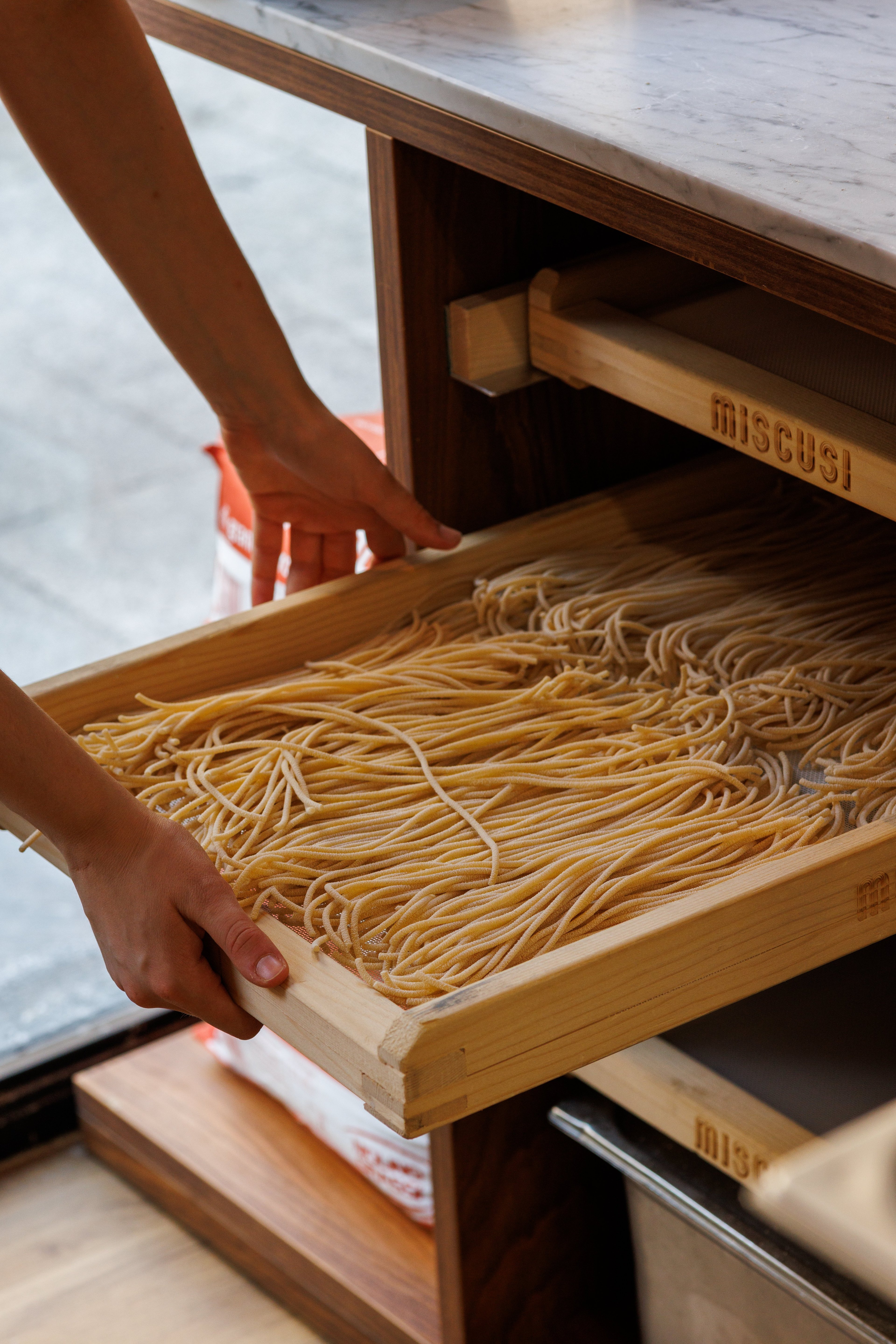 A person carefully slides a wooden tray filled with fresh, handmade pasta into a storage unit.